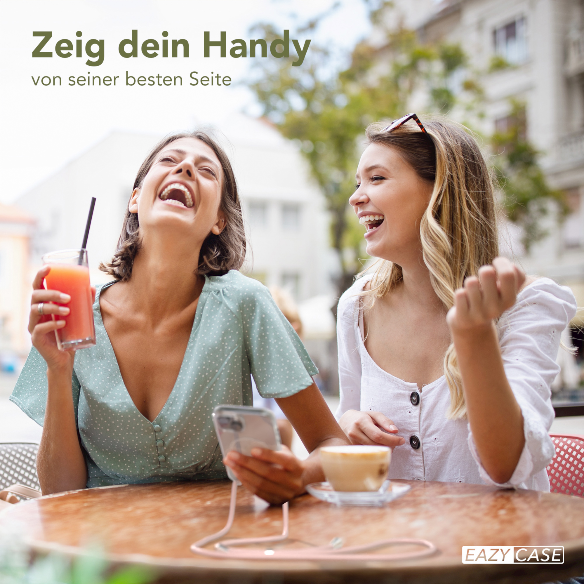 Zwei Frauen lachen draußen. Eine hält ein Getränk, die andere ein Handy, am Tisch mit Tasse.