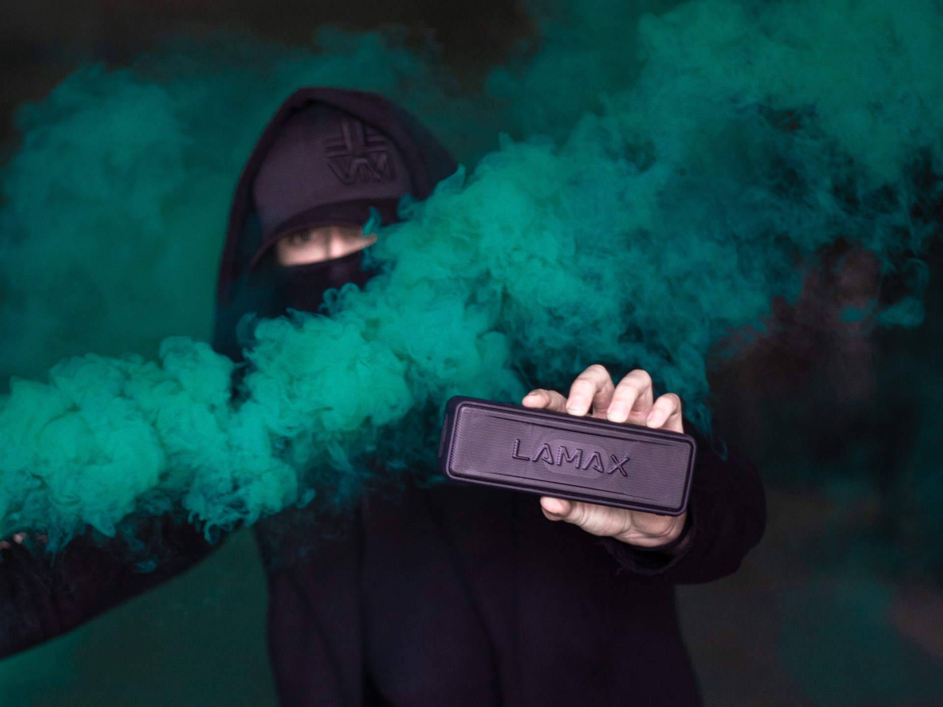 Una persona con una felpa nera con cappuccio tiene in mano un altoparlante LAMAX nero, con fumo verde.
