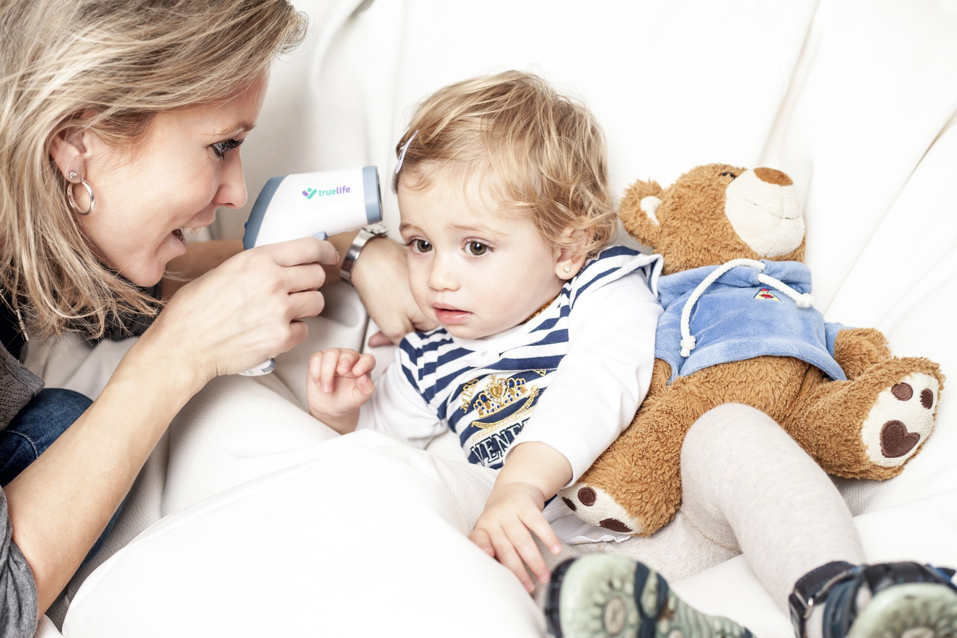 Frau misst Kind Temperatur mit Thermometer. Kind sitzt mit Teddybär.