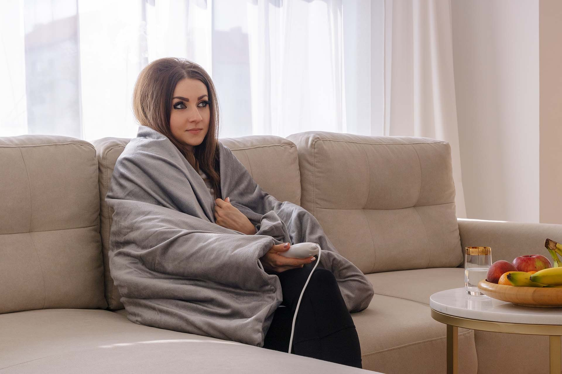 Kobieta owinięta w szary koc na beżowej sofie, trzymająca pilota. Owoce i woda na stole.