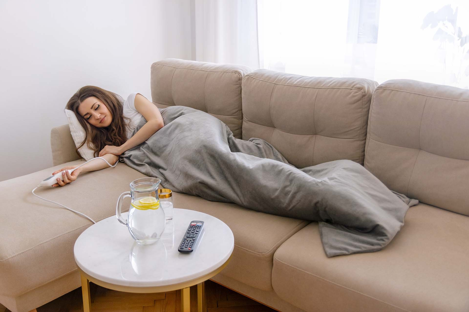 Kobieta śpiąca na beżowej sofie pod szarym kocem elektrycznym, z pilotem i wodą.