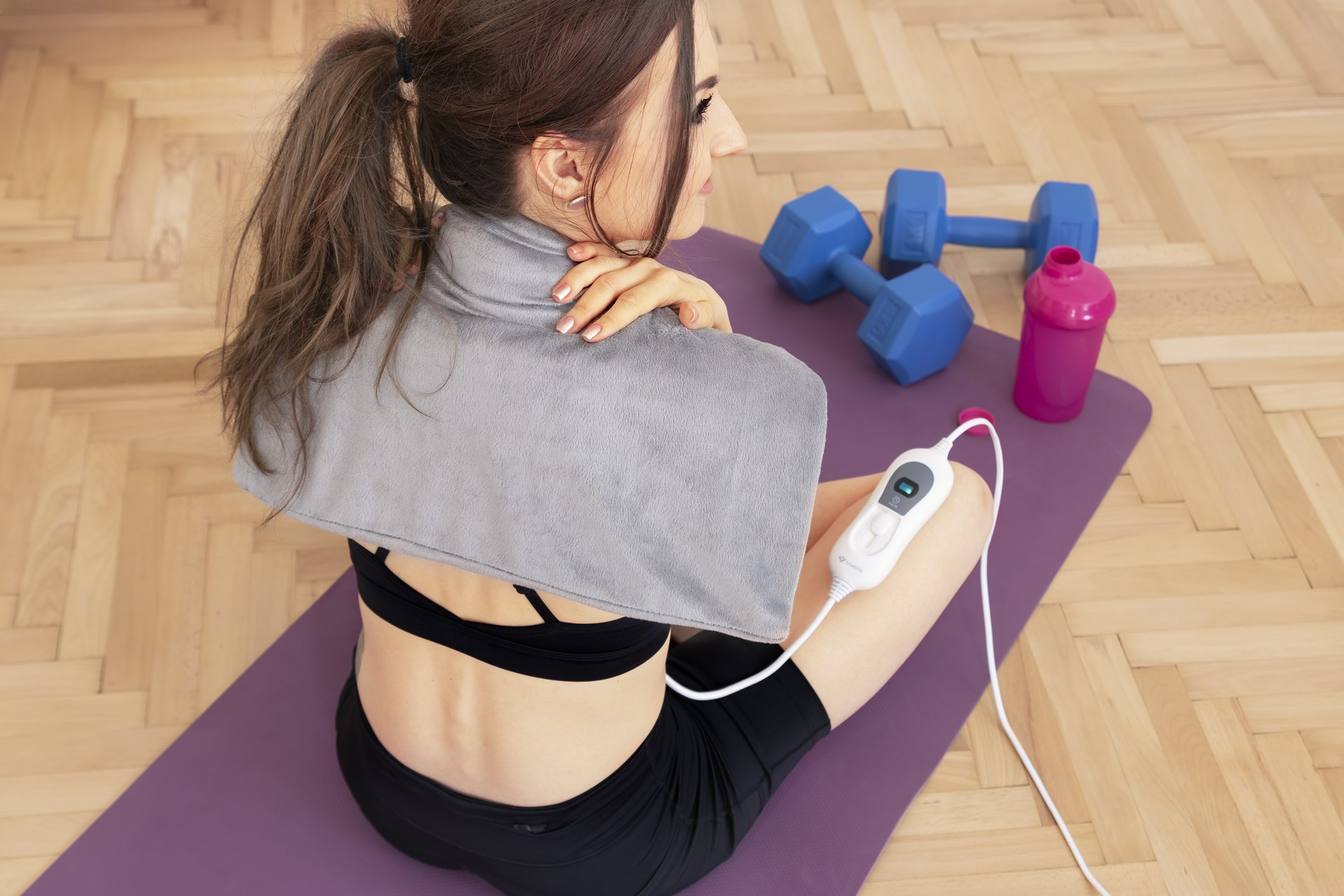 Kobieta z opaską na szyję na macie, hantlami i butelką wody. Drewniana podłoga.
