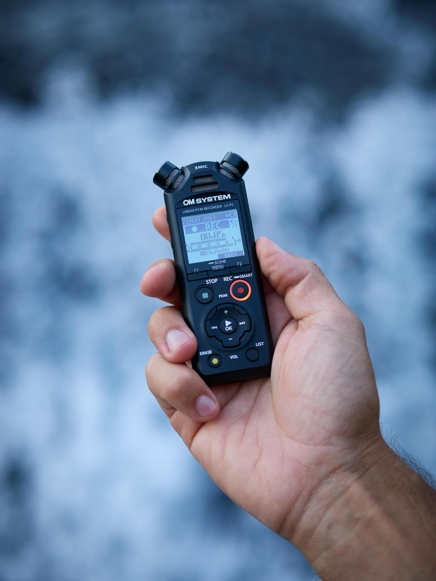 Hand hält schwarzen Audiorecorder, Bildschirm zeigt Informationen, verschwommenes Wasser im Hintergrund.