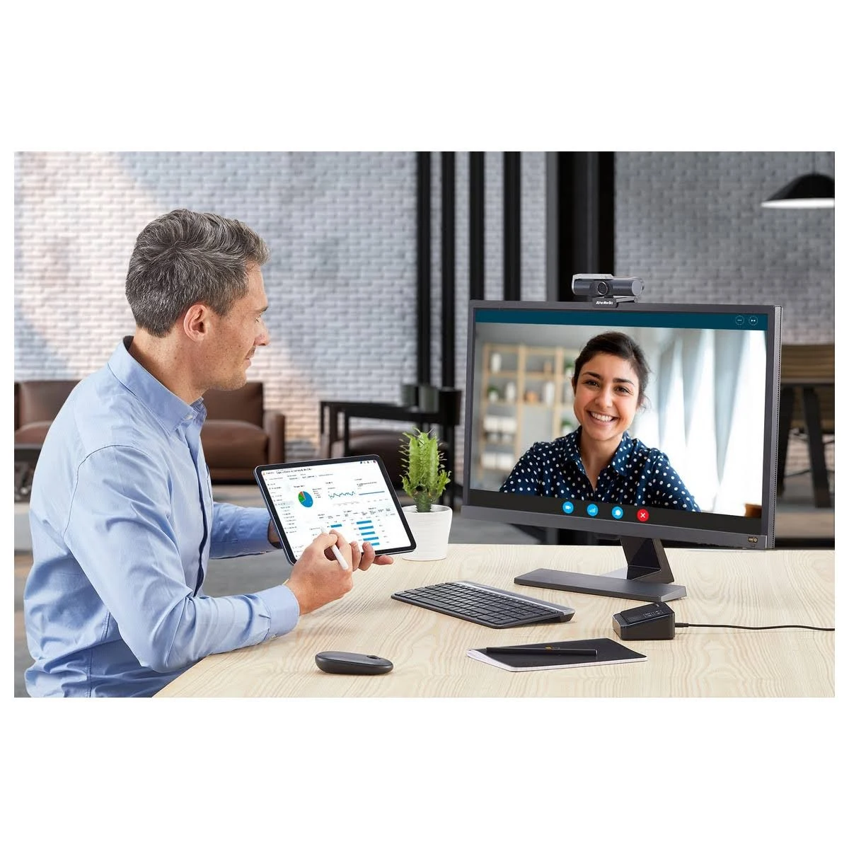 Mężczyzna używający tabletu i kobieta na ekranie monitora. Pokój zawiera klawiaturę, mysz i roślinę.