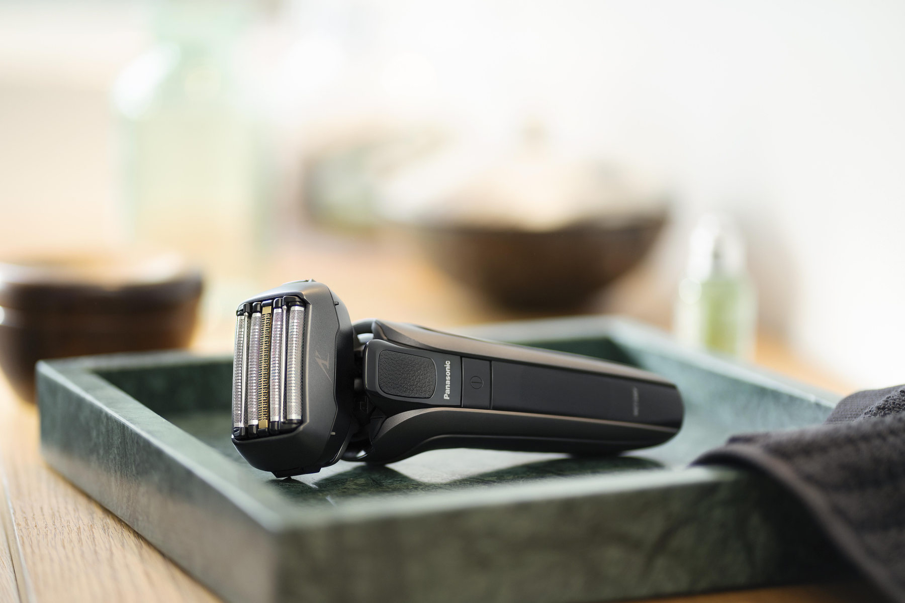 Czarna i srebrna golarka elektryczna na zielonej tacy. Butelka i słoik w tle.