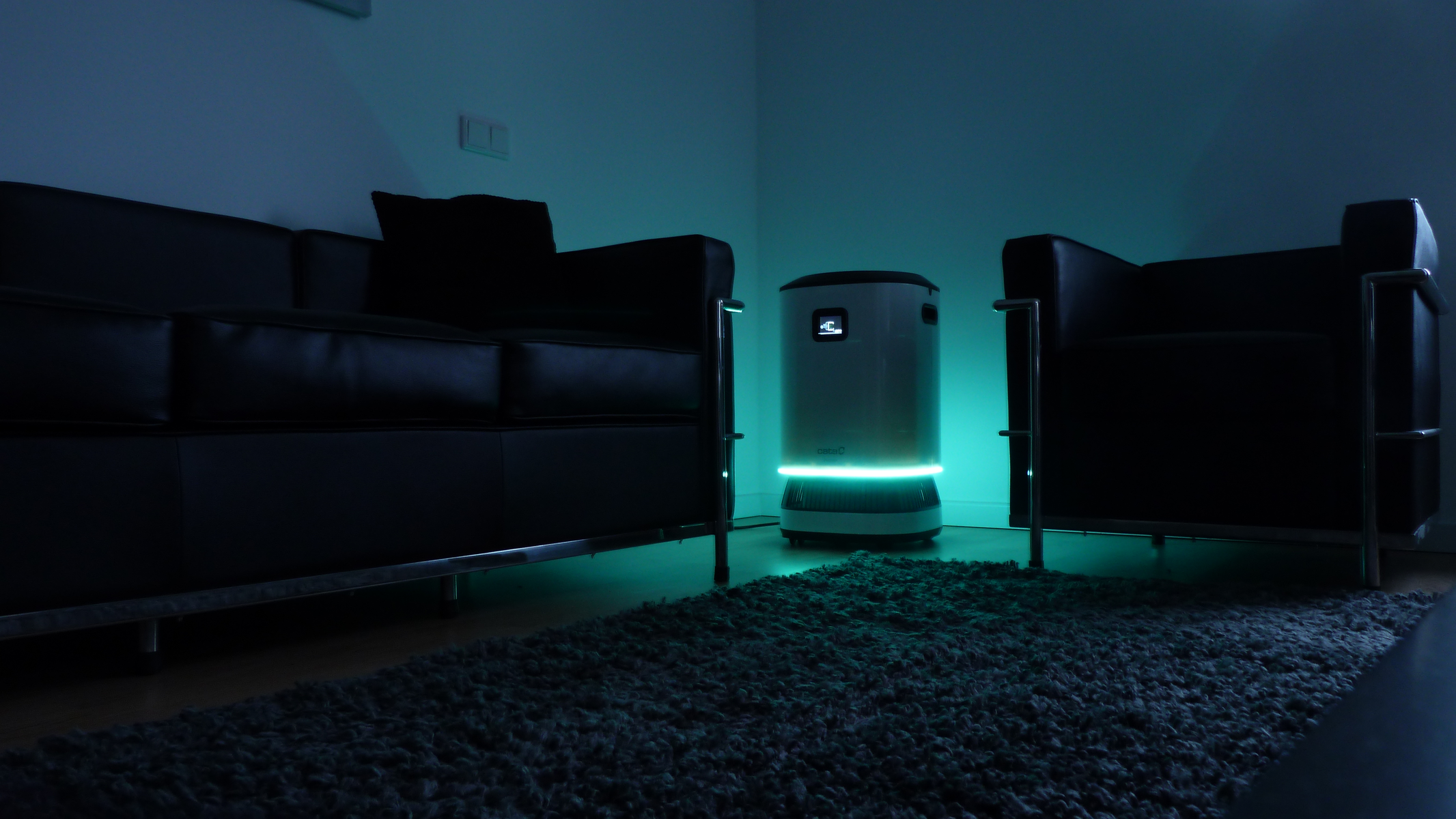 Un robot blanco se sienta entre dos sillas negras, en una habitación con luces verdes. Hay una alfombra.