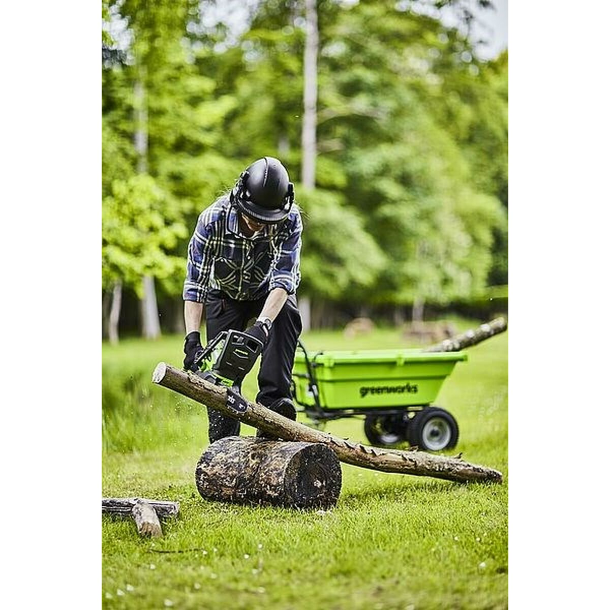 Person mit Kettensäge an einem Baumstamm auf einer Wiese. Greenworks-Kettensäge.