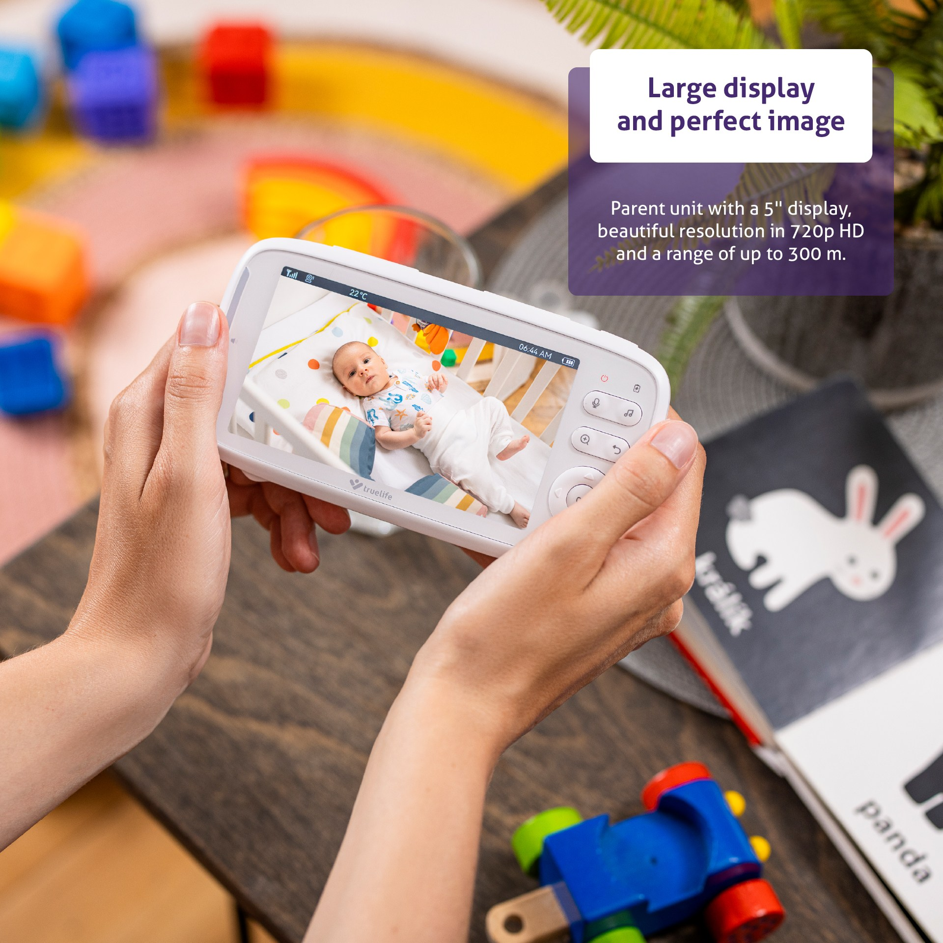 Hände, die einen Babyphone halten, das ein Baby zeigt. Spielzeug und ein Pandabuch sind in der Nähe.