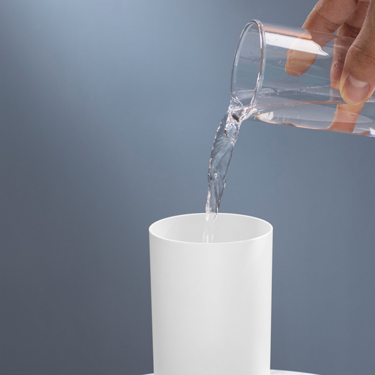 Hand gießt Wasser aus einem Glas in eine weiße Tasse. Blau-grauer Hintergrund.