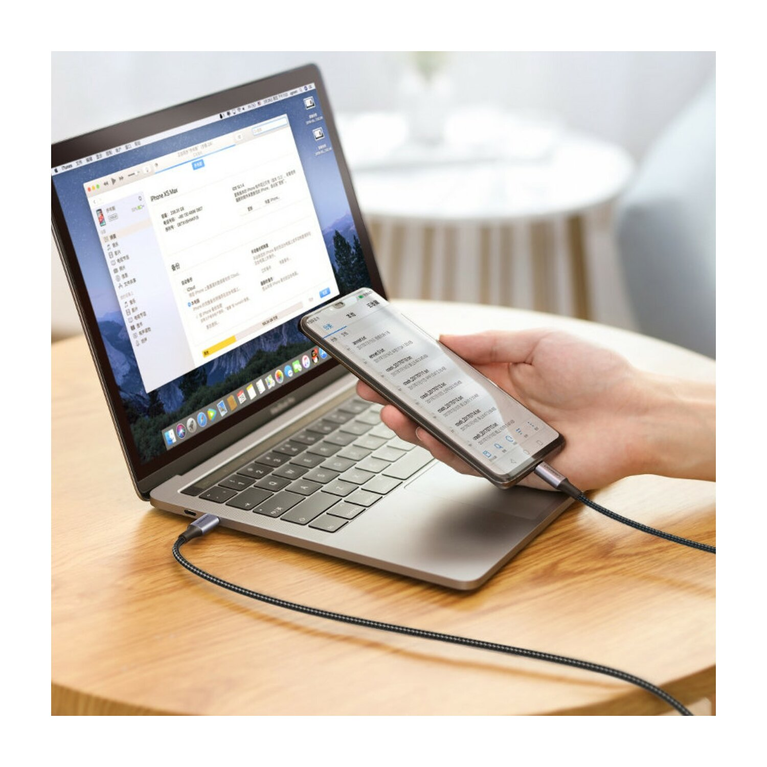 Dłoń trzymająca telefon podłączony do laptopa kablem. Laptop stoi na drewnianym stole.