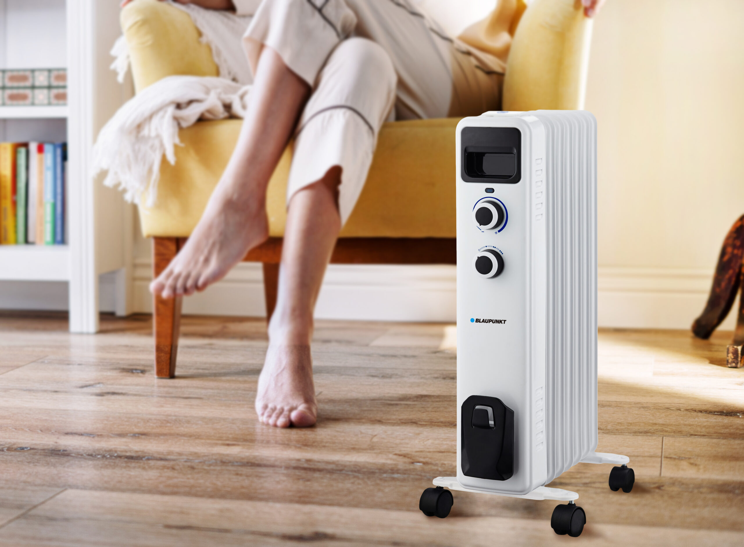 Biały grzejnik olejowy na kółkach stoi blisko osoby siedzącej na żółtym krześle. Podłoga jest drewniana.
