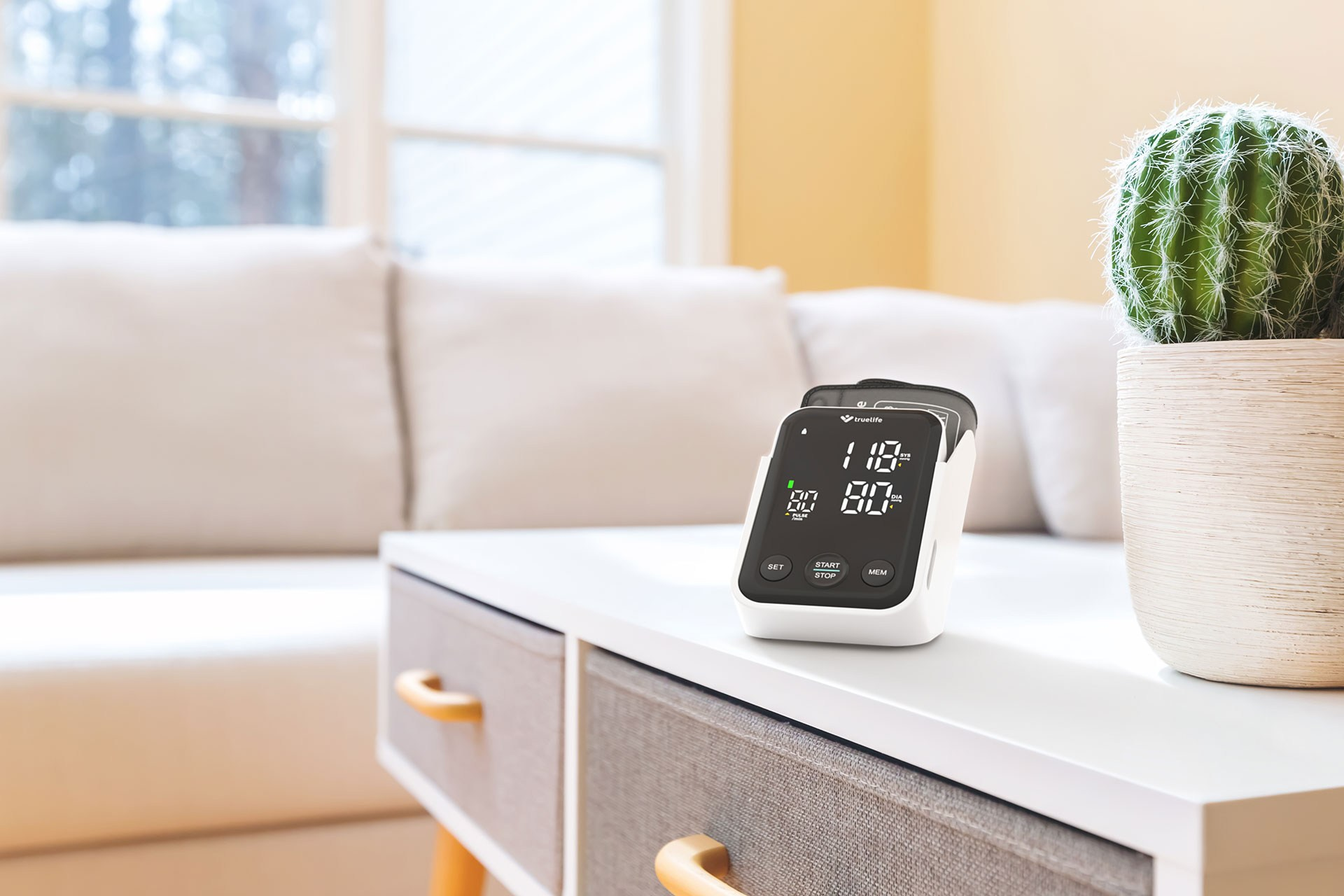 Biały monitor ciśnienia krwi na białym stole, z kaktusem w doniczce w tle.