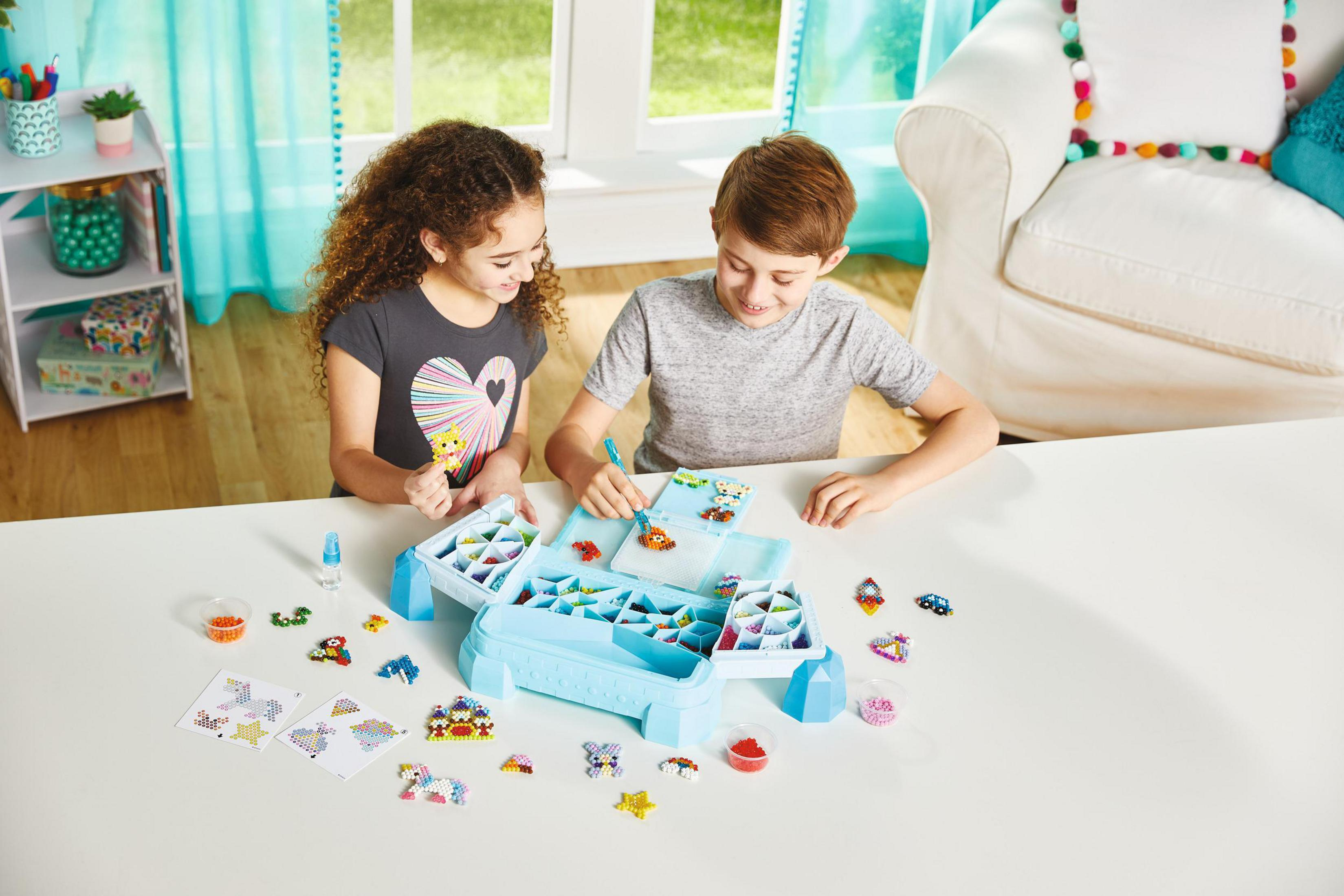 Due bambini giocano con un set di perline. Una scatola blu con perline e disegni è visibile.