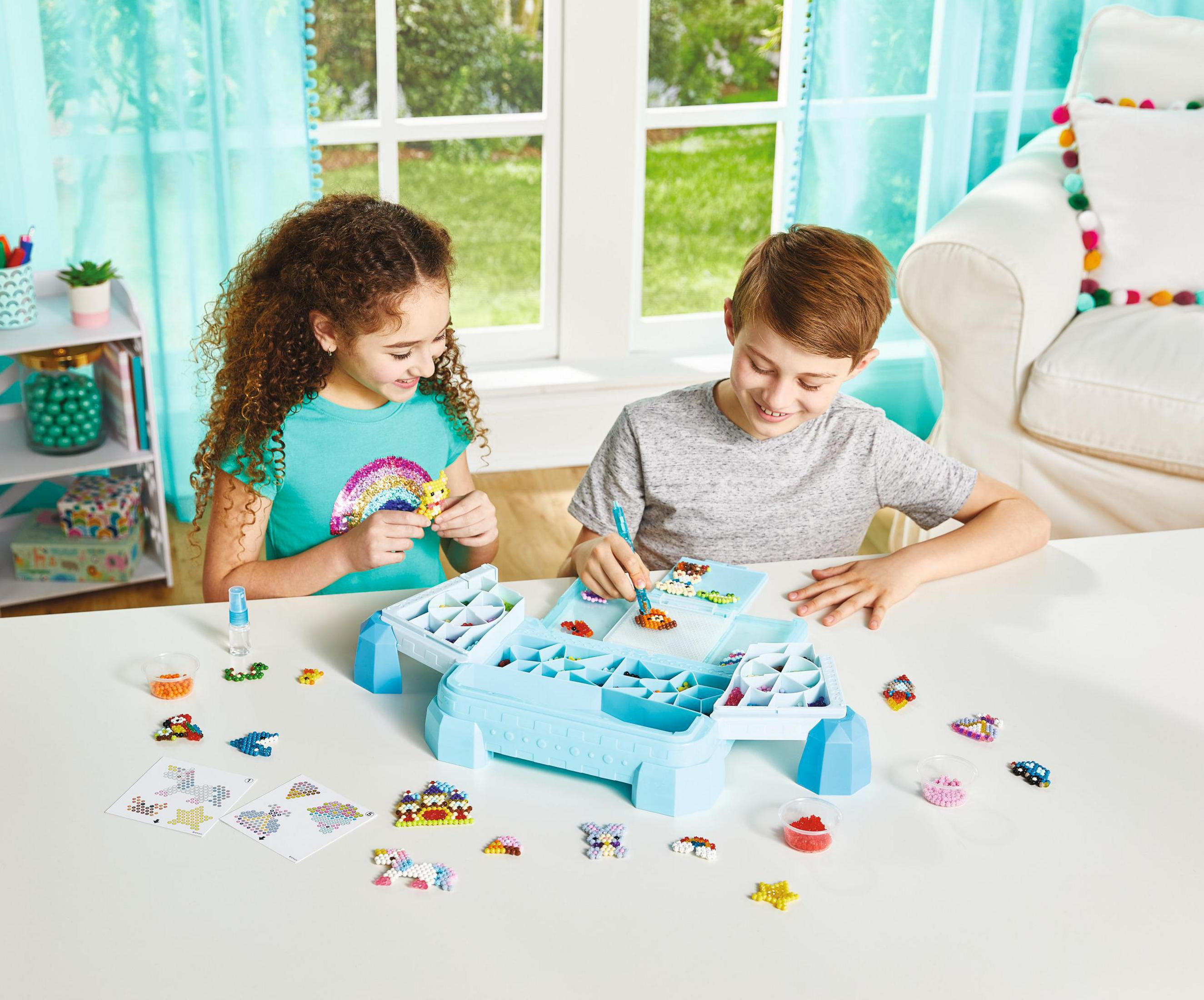 Due bambini che creano con perline, organizer blu, tavolo bianco e vista finestra.