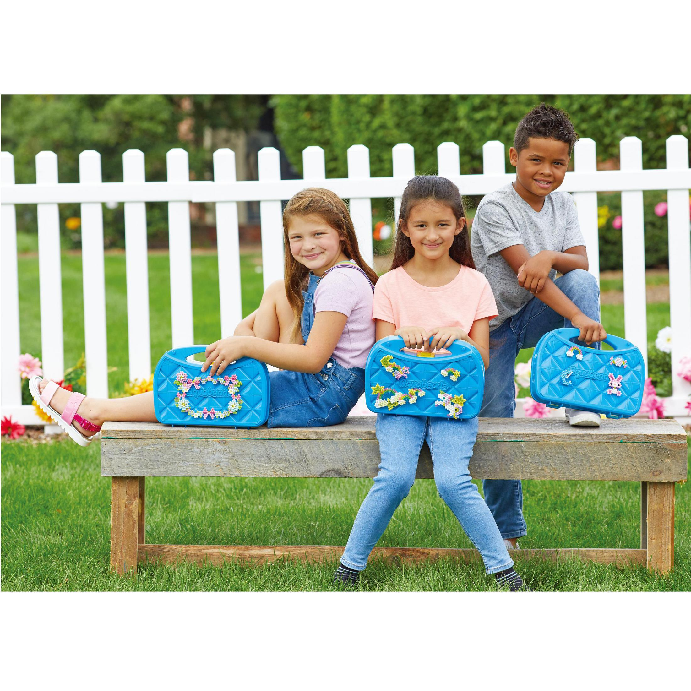 Drei Kinder sitzen auf einer Holzbank und halten blaue Taschen mit Blumendekorationen.