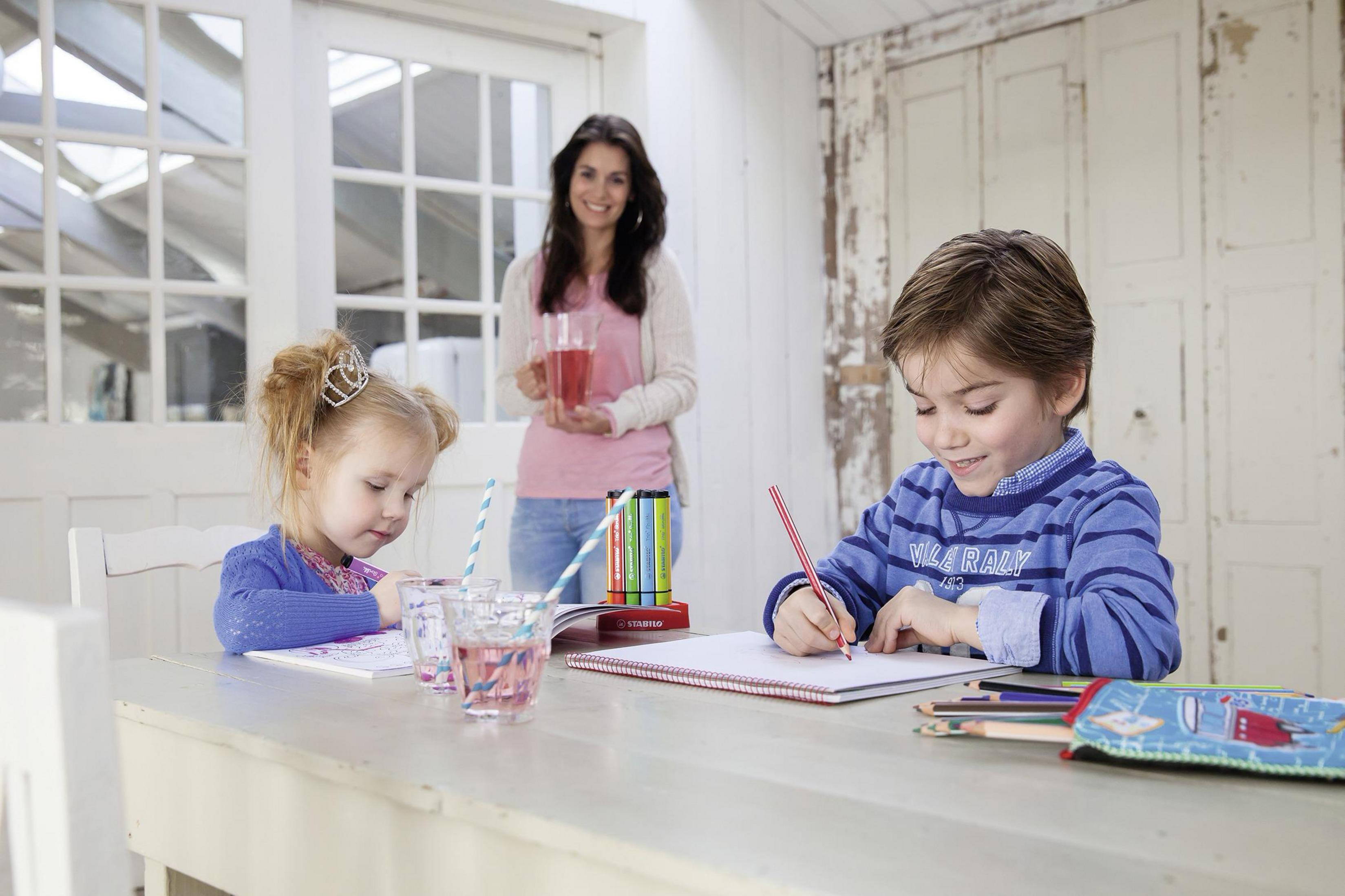 Zwei Kinder zeichnen am Tisch, eine Frau mit Getränk steht im Hintergrund.