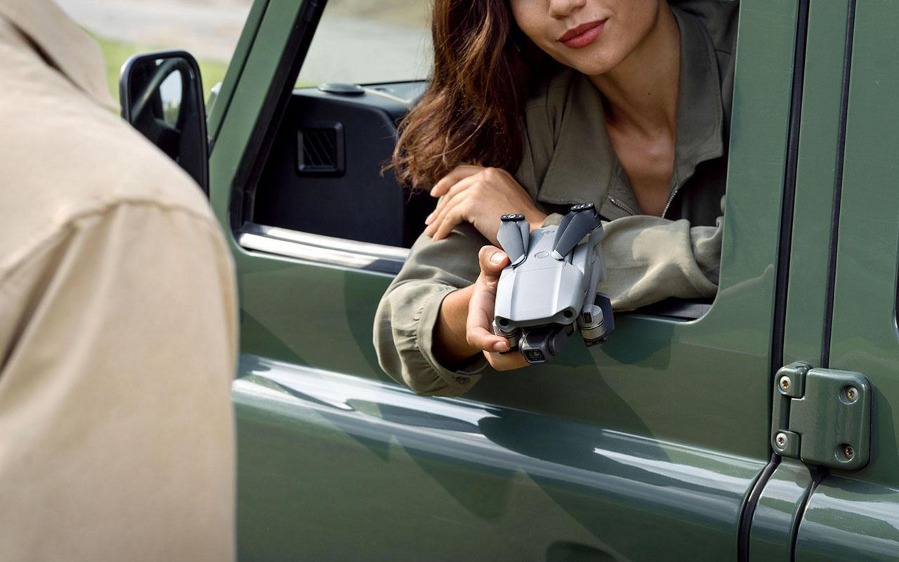 Eine Person gibt eine Drohne an eine andere Person, neben einem grünen SUV, die Frau hält die Drohne.