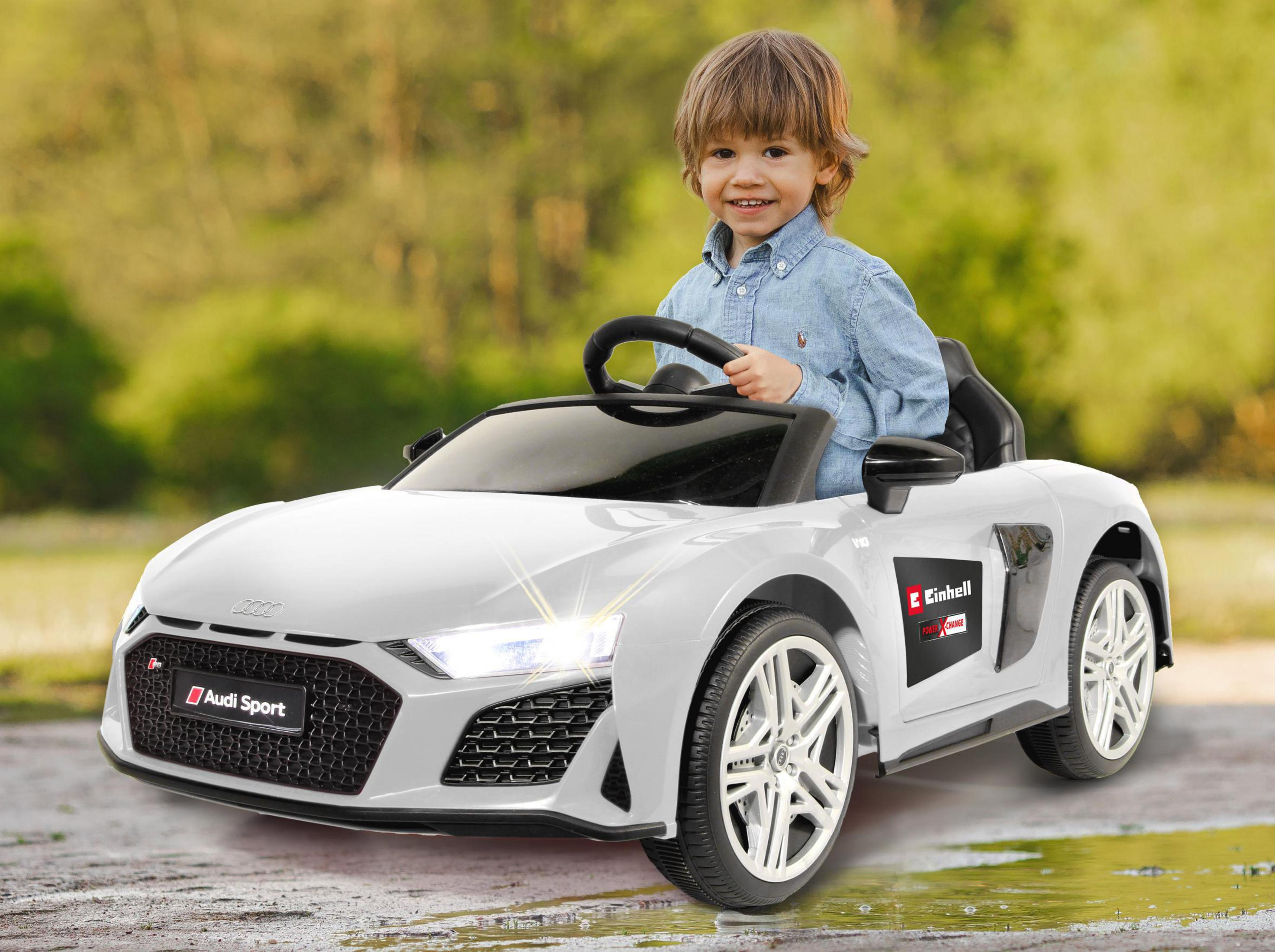 Ein Junge in einem weißen Spielzeugauto. Er lächelt. Das Auto hat Audi Sport-Branding und steht auf einer Straße.