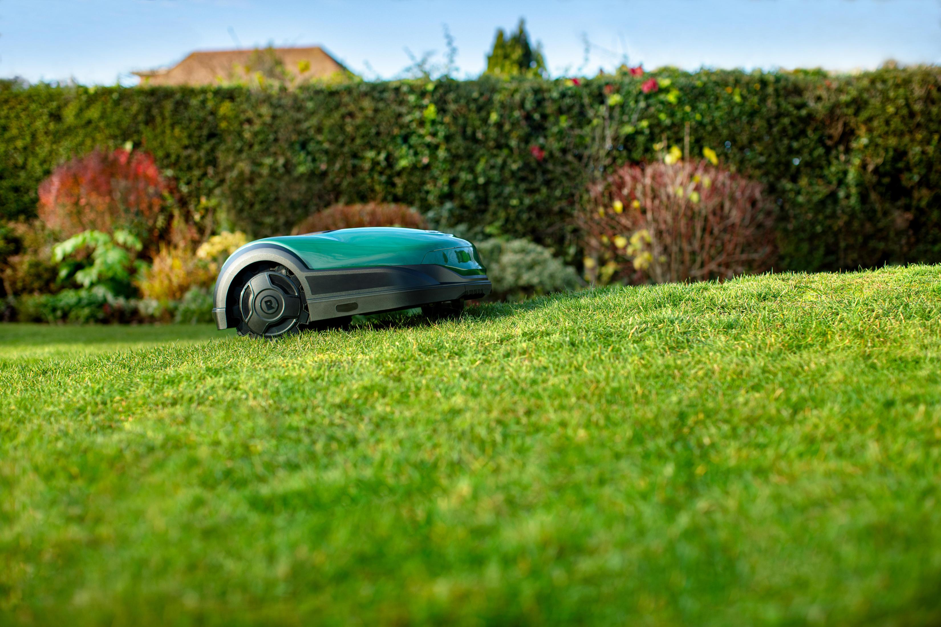 Zielona kosiarka robotyczna na pochyłym trawniku, z żywopłotem i drzewami w tle.