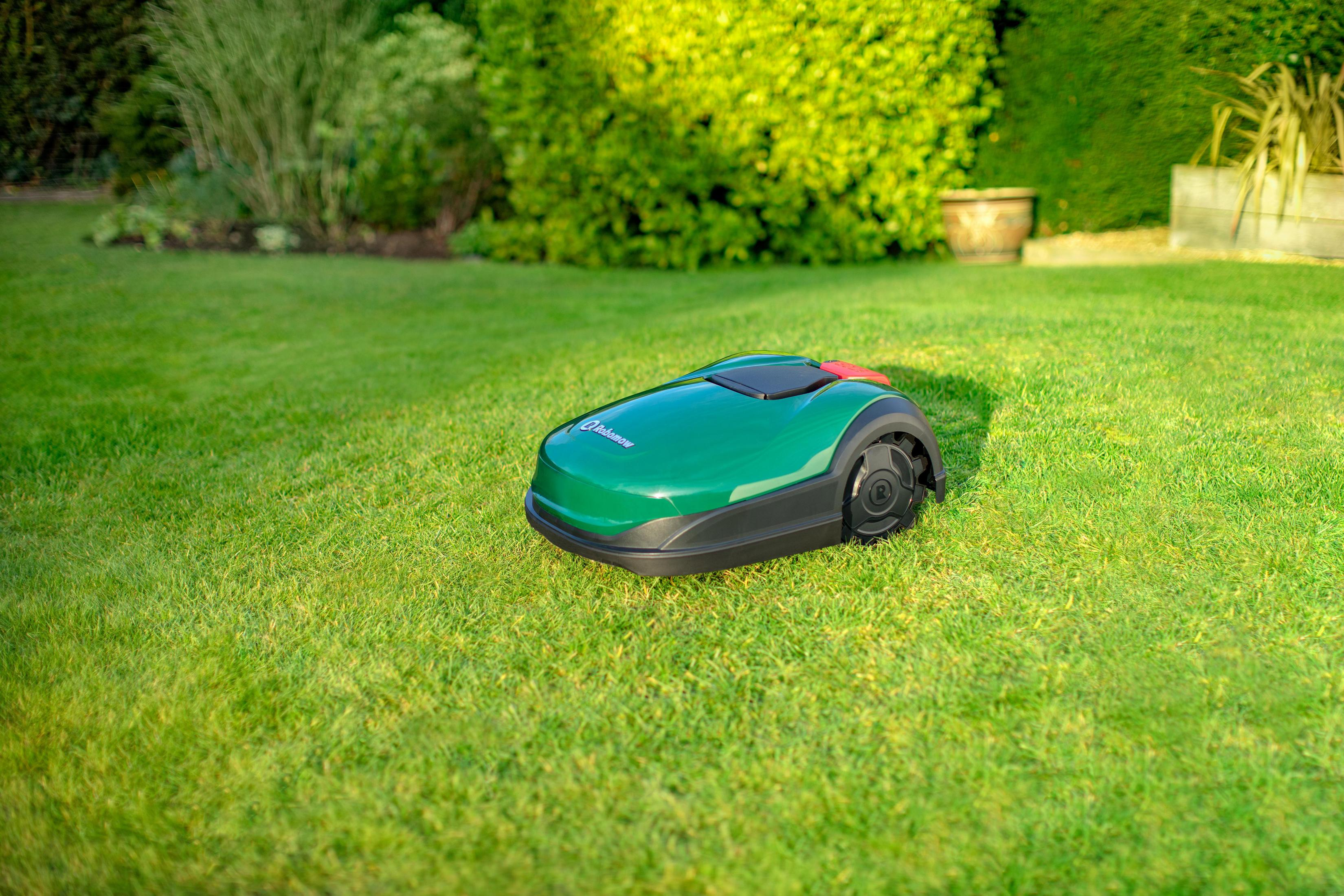 Zielona kosiarka robotyczna na zielonym trawniku, z zielonym żywopłotem w tle.