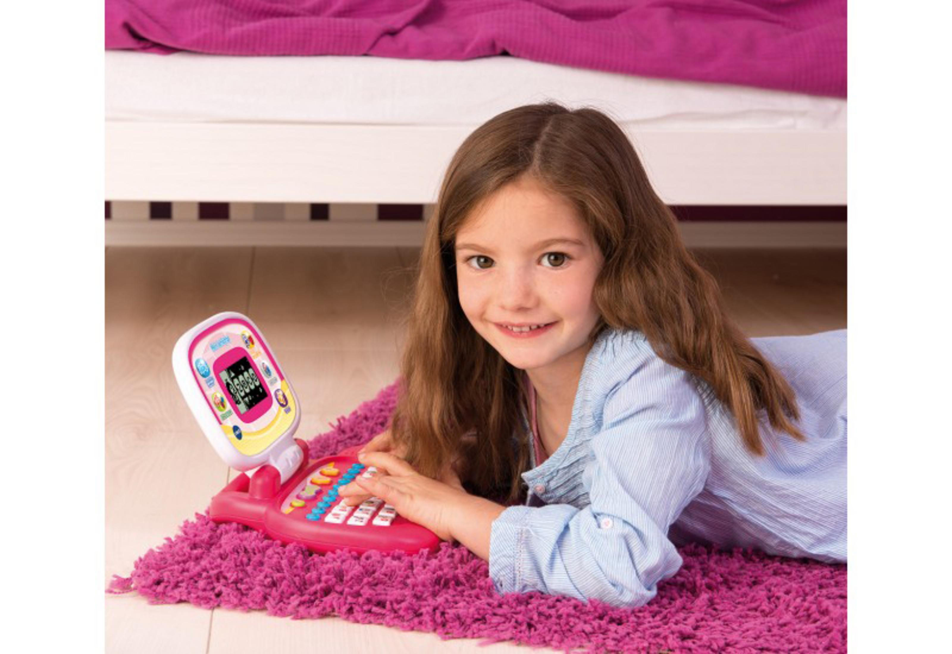 Ein junges Mädchen lächelt, während sie mit einem rosa Spielzeugtelefon auf einem rosa Teppich in der Nähe eines Bettes spielt.