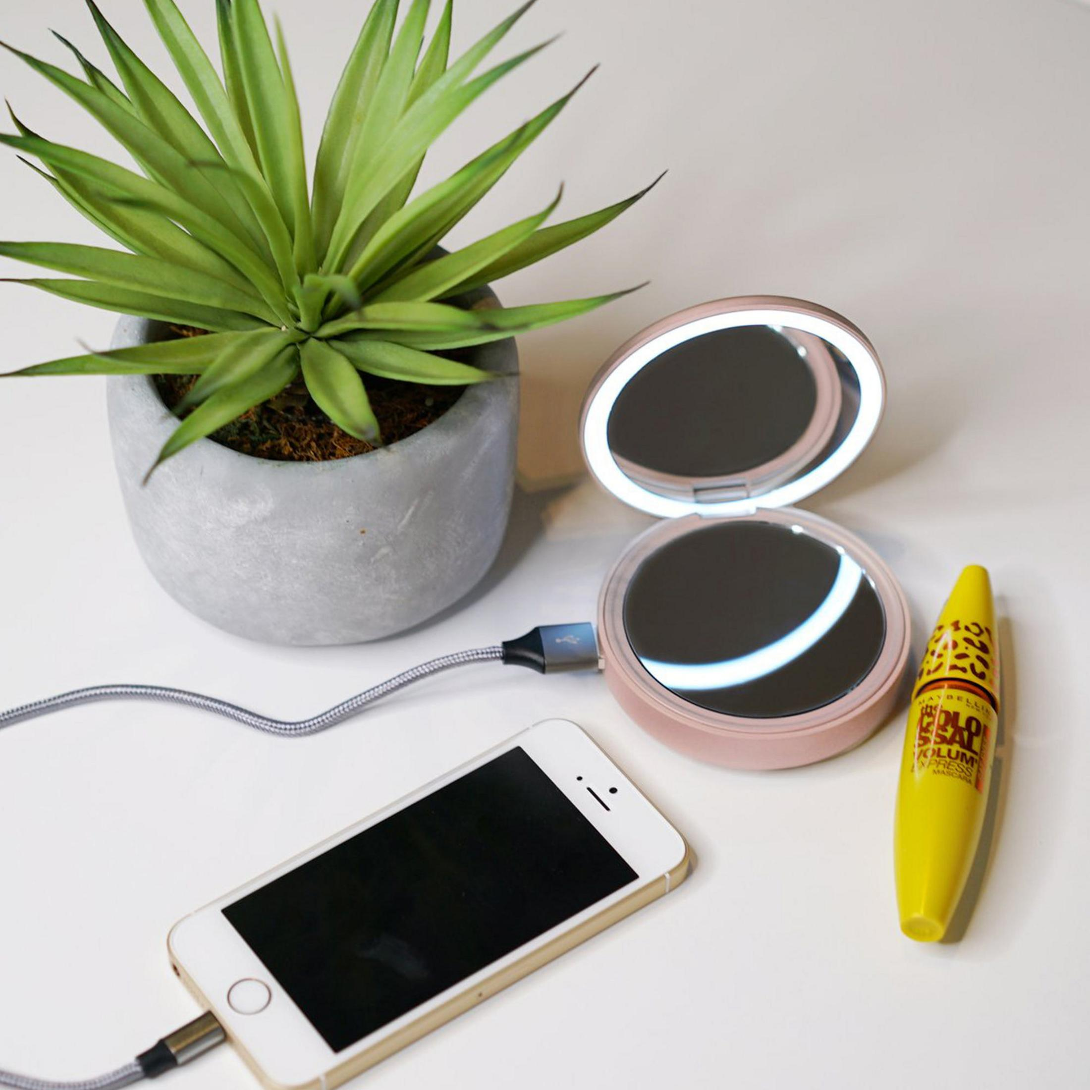 Ein Telefon, das in der Nähe eines Spiegels geladen wird, eine kleine Pflanze und eine Mascararöhre auf einem weißen Tisch.