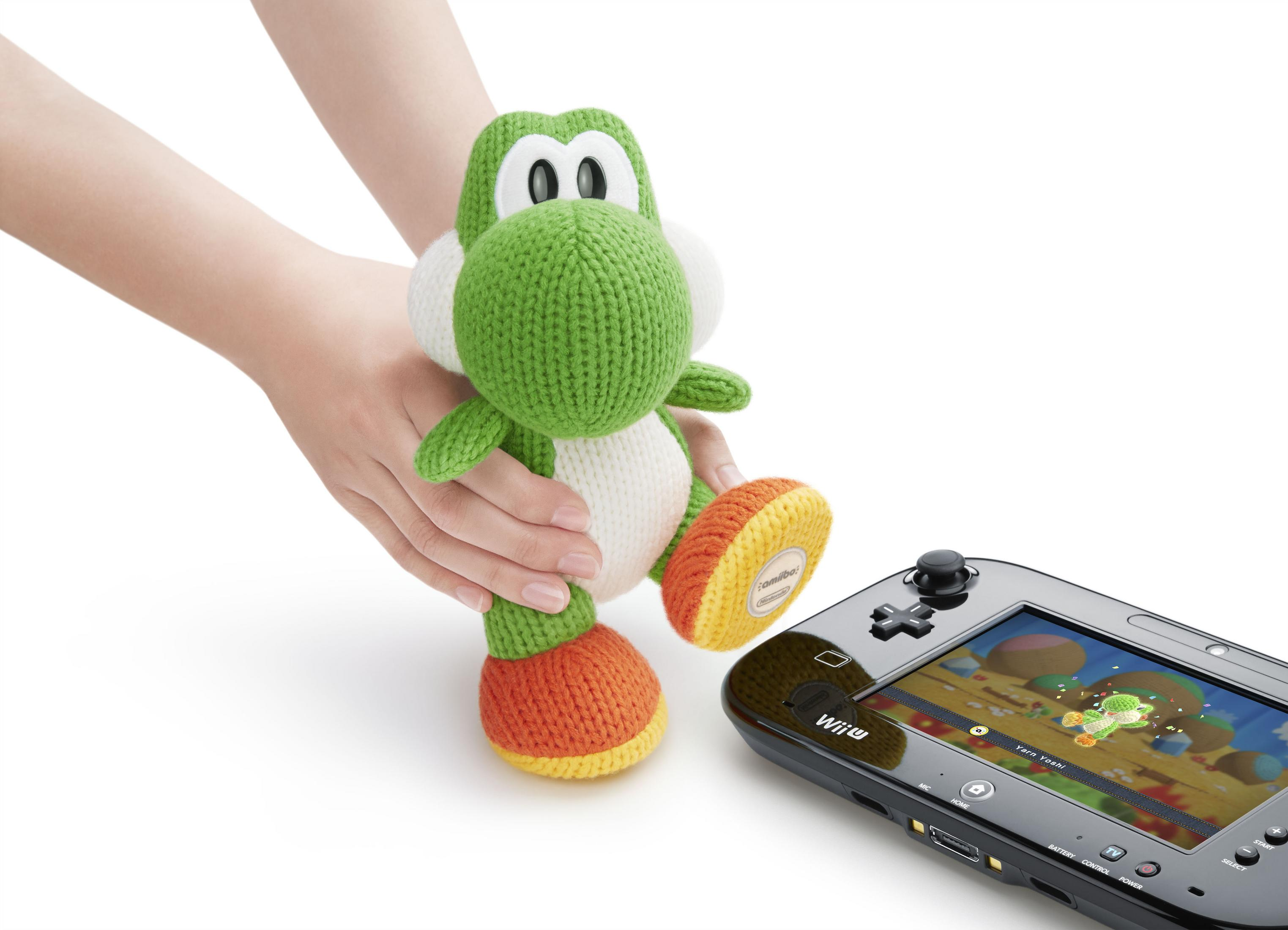 Eine Person hält ein gestricktes Yoshi-Spielzeug neben einer Wii U-Konsole, die einen Spielbildschirm anzeigt.