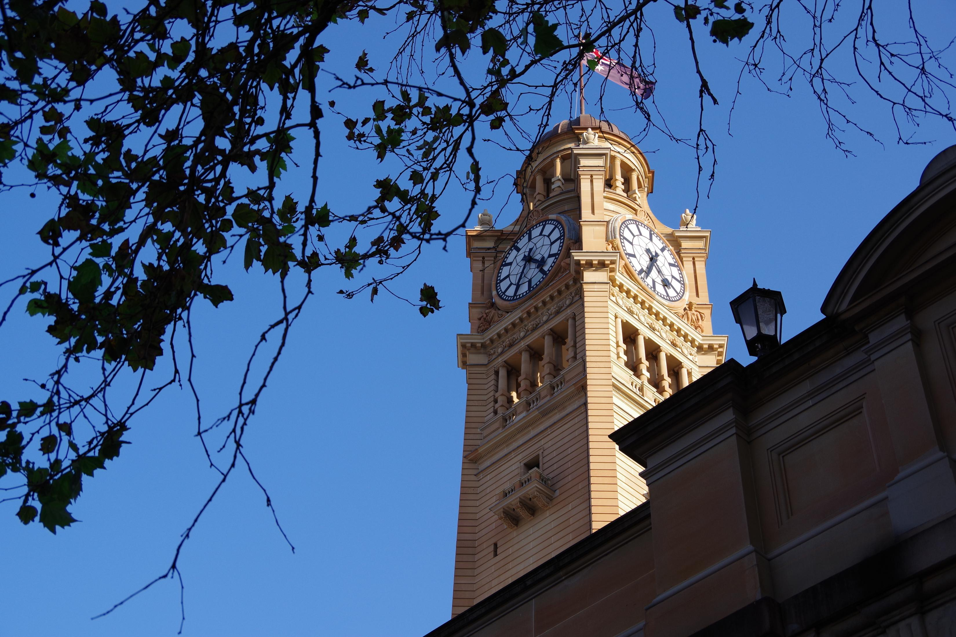 Ein Uhrturm mit zwei Zifferblättern, von unten gesehen. Ein blauer Himmel und Äste sind sichtbar.