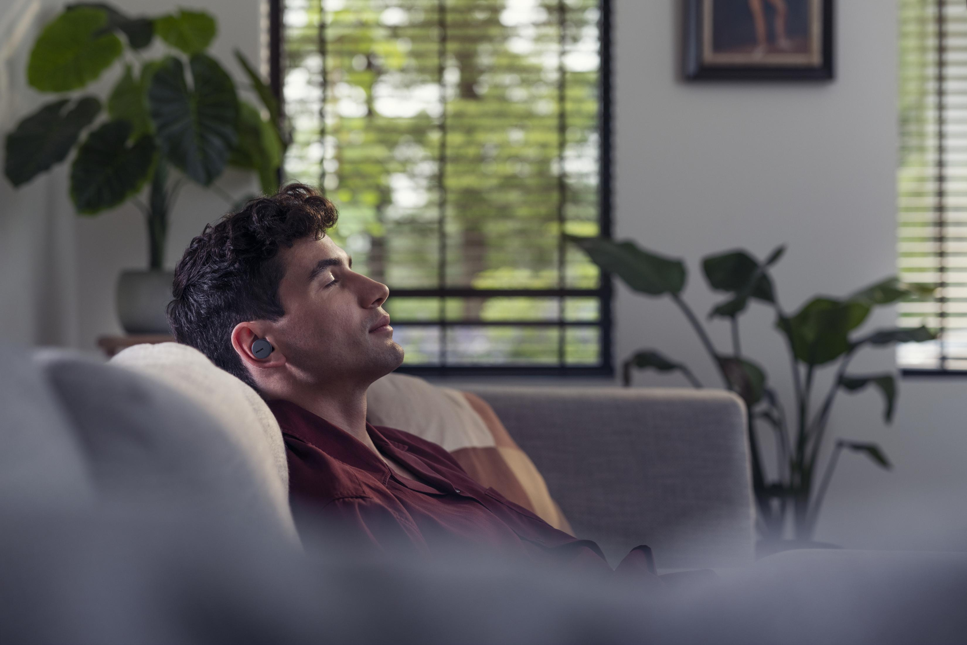 Ein Mann sitzt auf einem Sofa mit Ohrhörern, Augen geschlossen, in einem gut beleuchteten Raum.