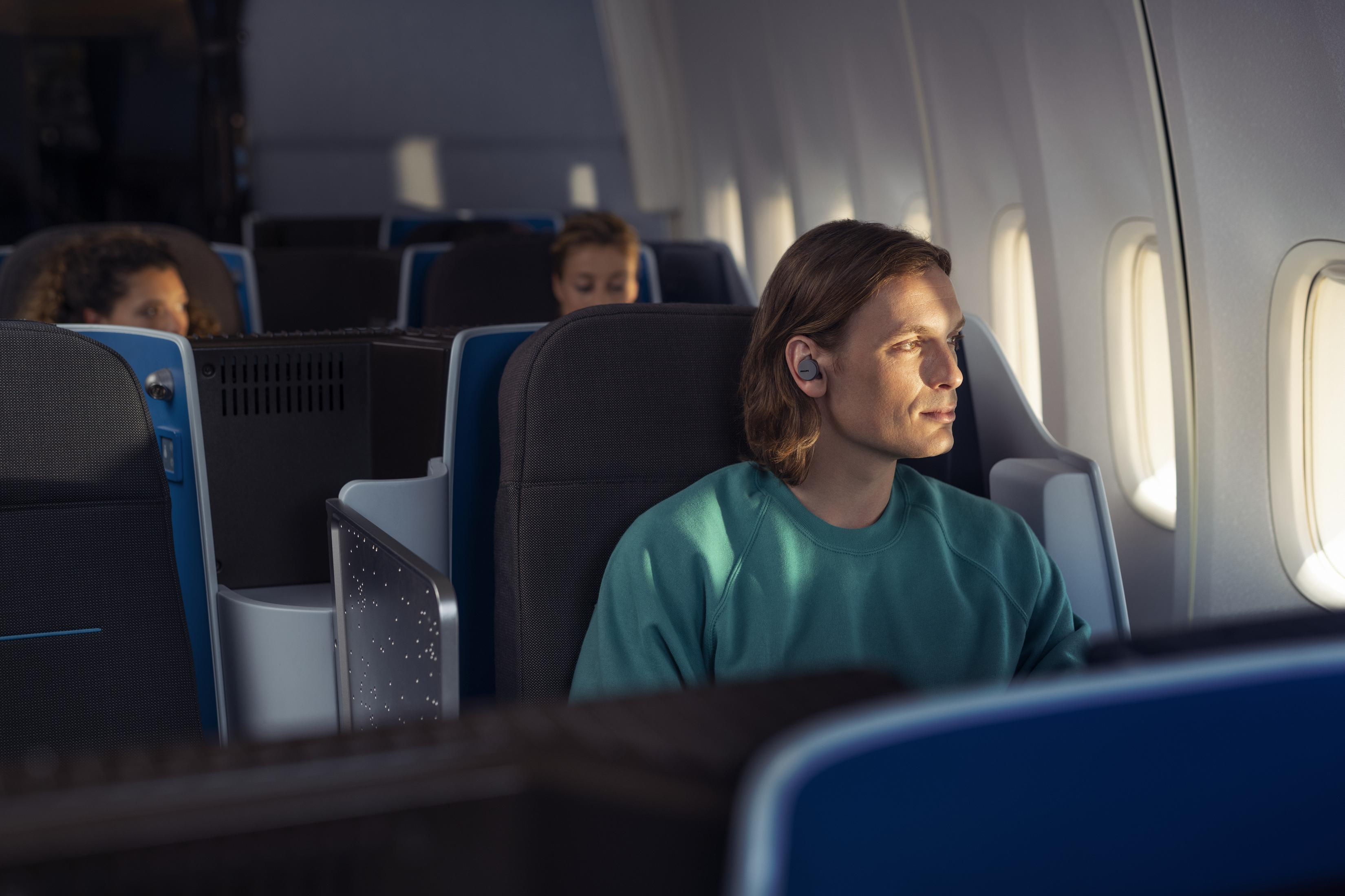 Mann im Flugzeug mit Ohrhörern, schaut aus dem Fenster. Andere Passagiere im Hintergrund.