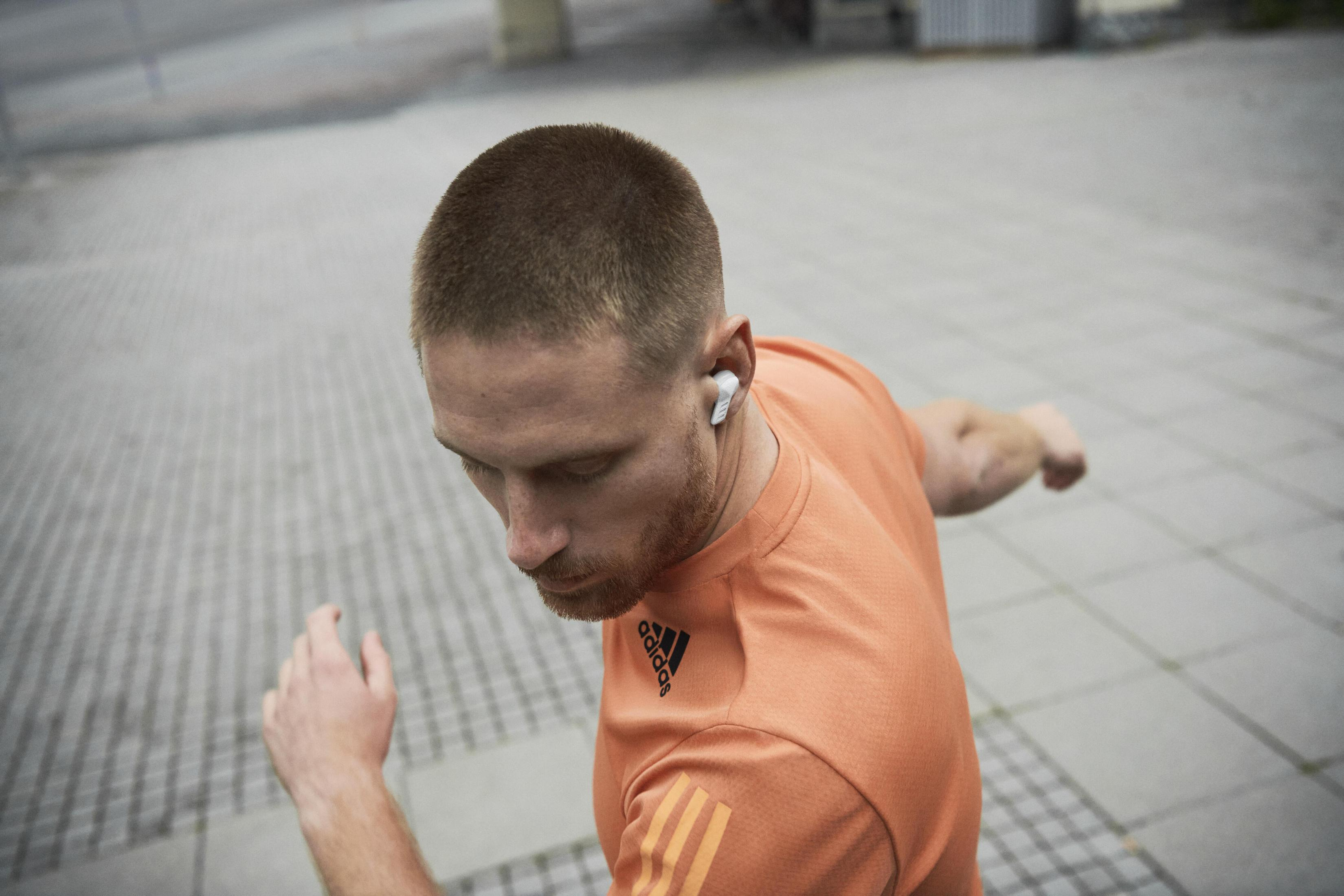 Ein Mann, der im Freien läuft, trägt ein orangefarbenes Adidas-Shirt und weiße Ohrhörer.