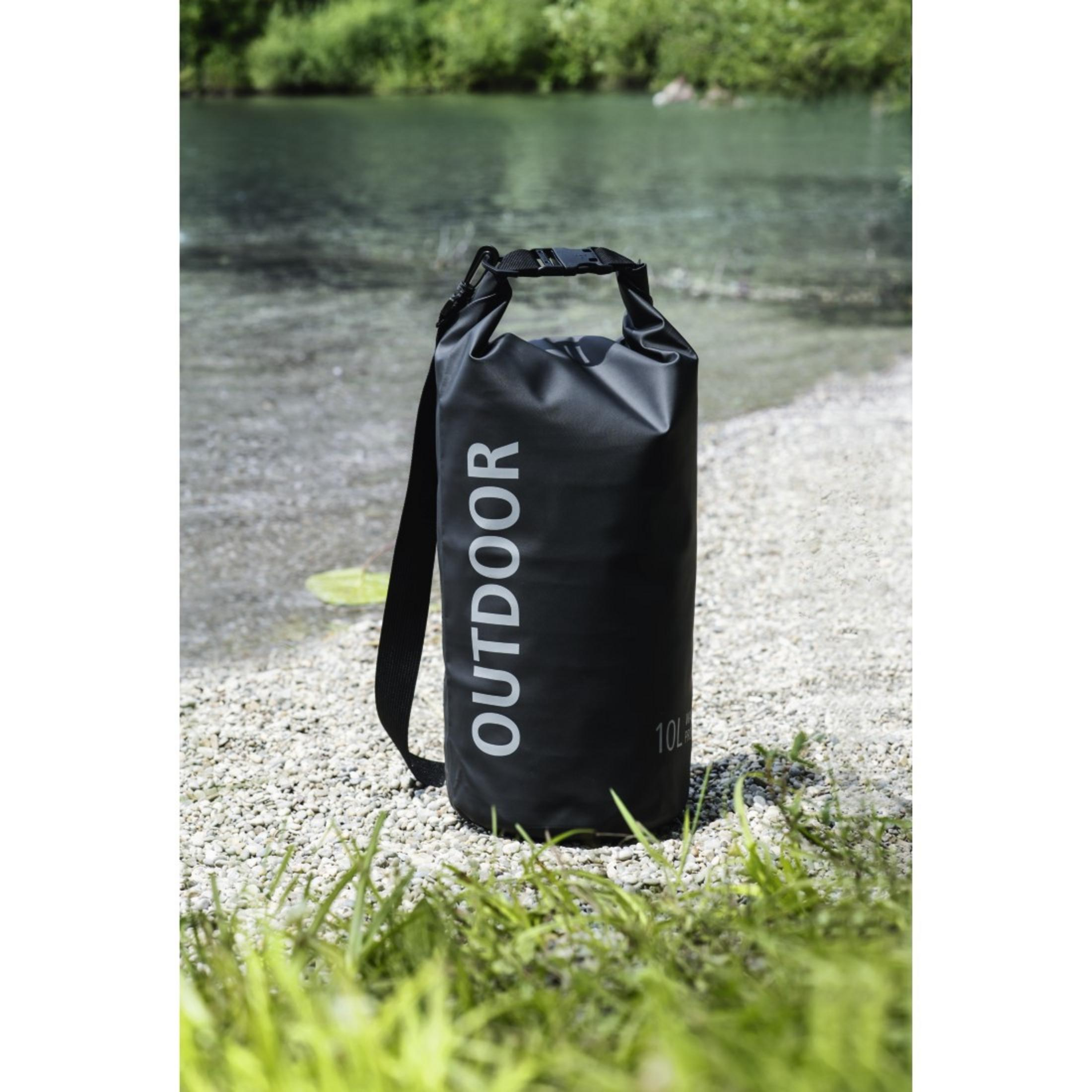 Eine schwarze Outdoor-Trockentasche auf einem Kiesstrand mit grünem Gras davor und Wasser und Bäumen im Hintergrund.