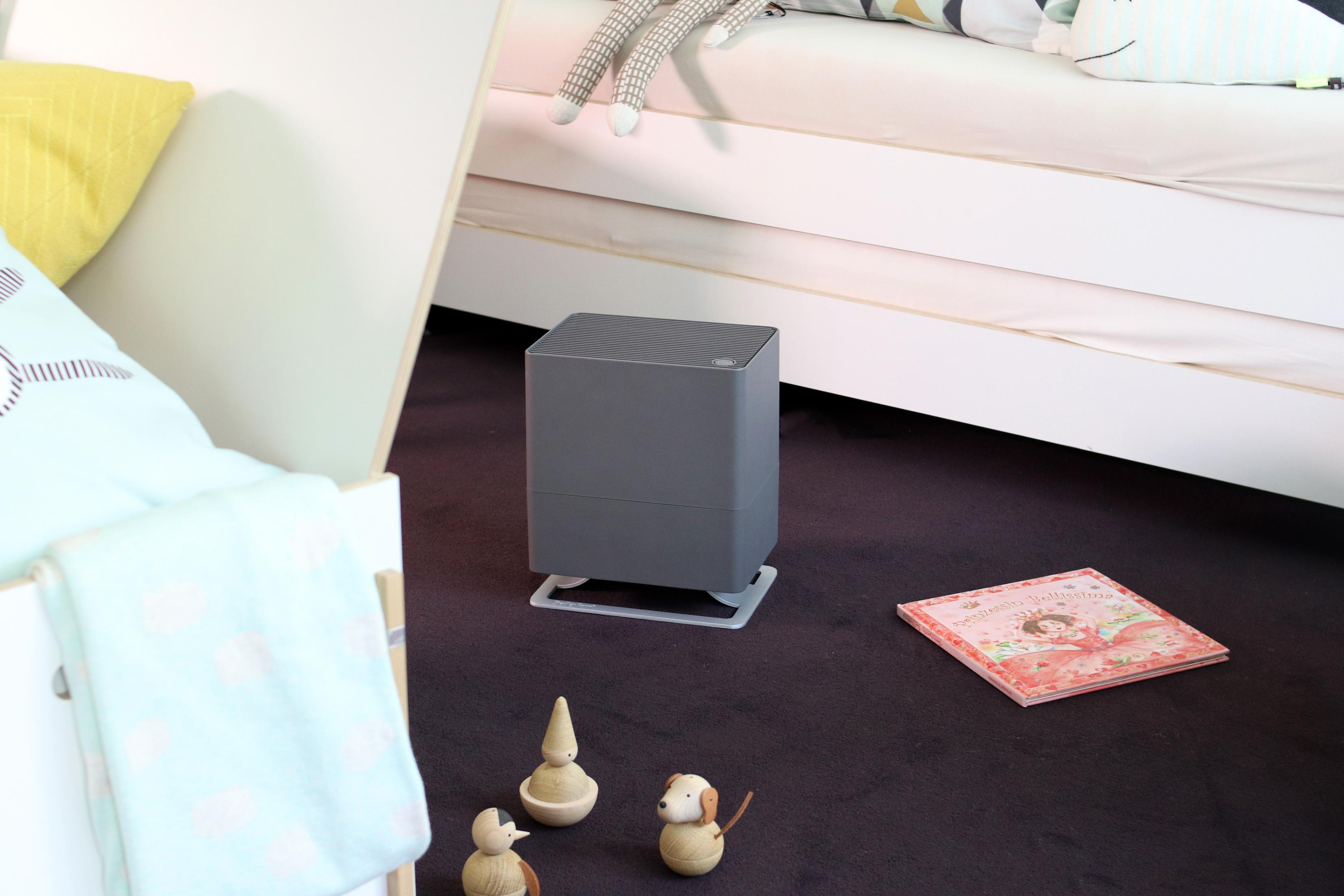 A gray humidifier on a dark carpet. A children's book and wooden toys are nearby.
