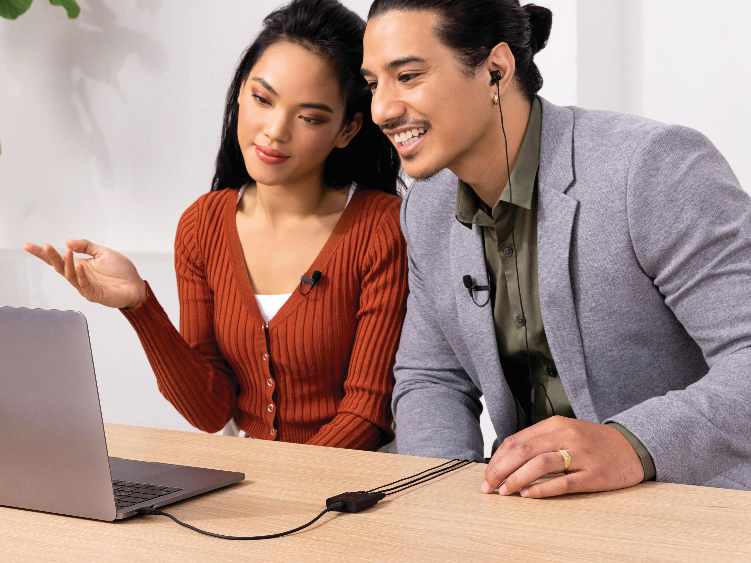 Twee mensen kijken naar een laptop. Beiden dragen microfoons. Ze zitten aan een houten tafel.