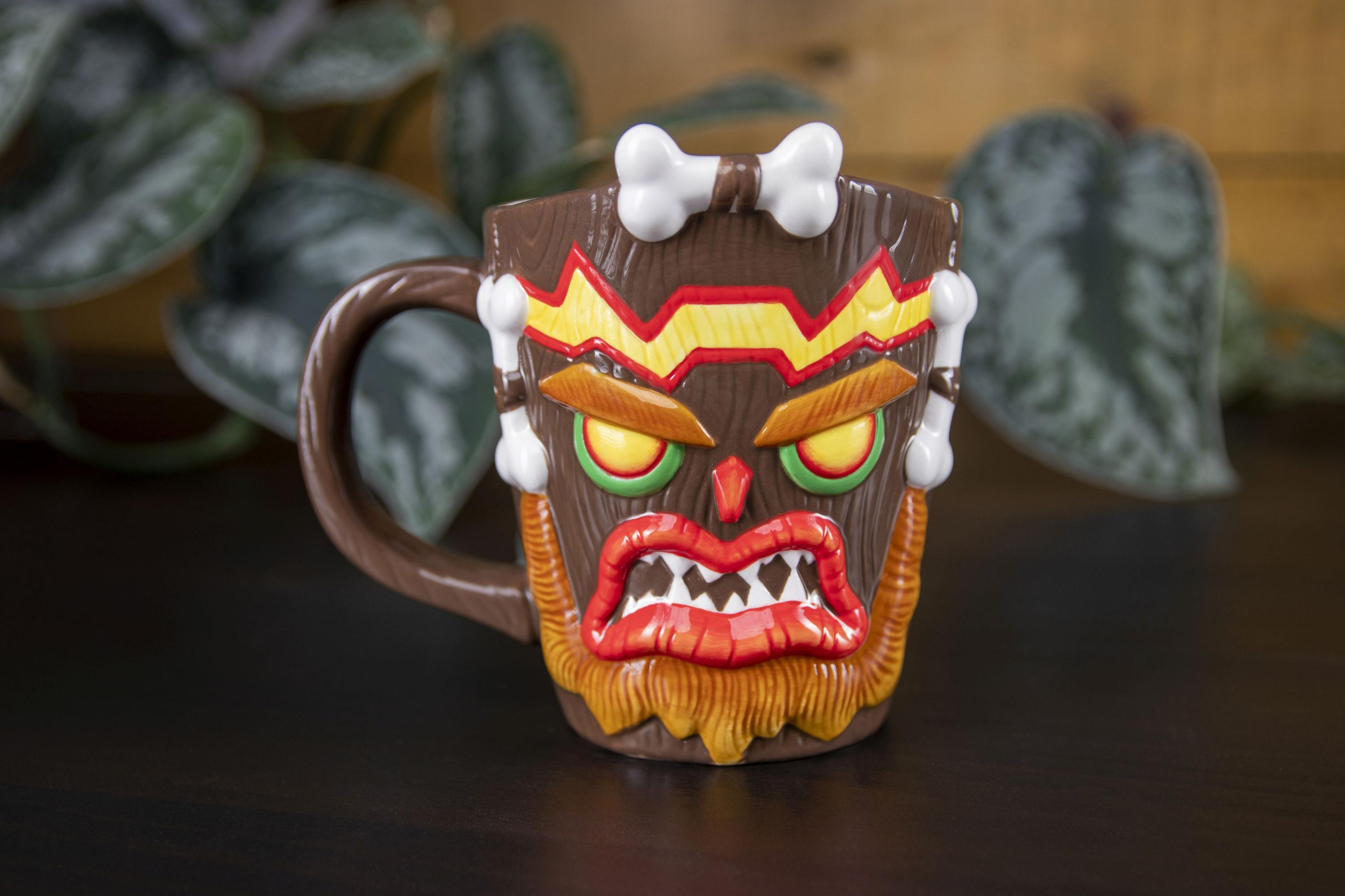 Eine braune Tiki-Tasse mit Knochenakzenten, grünen Augen und einem orangefarbenen Bart steht vor einer Pflanze.