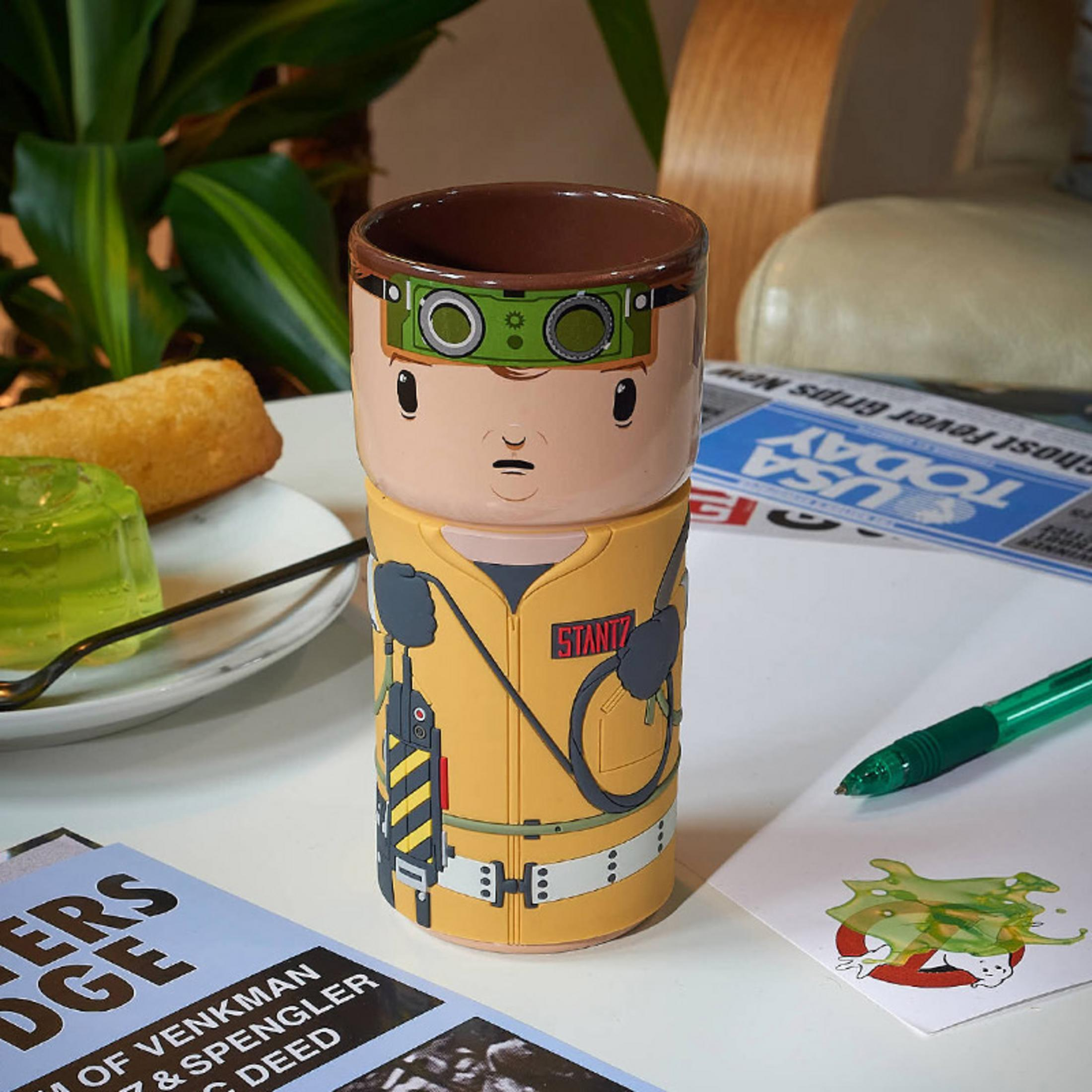 Ghostbusters-Tasse mit Cartoon-Figur, Essen und Papieren im Hintergrund.