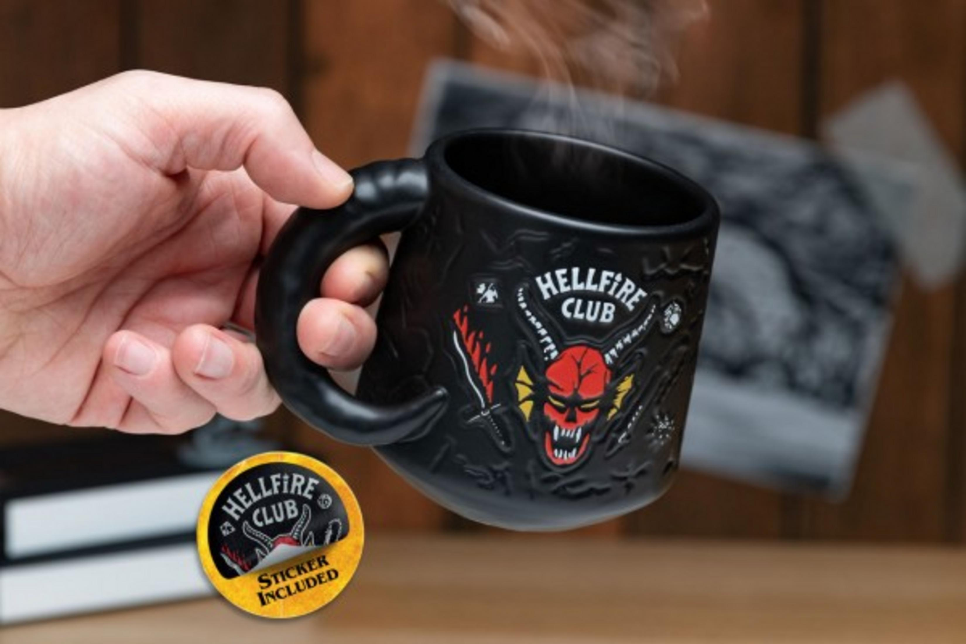 Schwarze Tasse mit Hellfire Club Logo und Aufkleber. Von Hand gehalten, auf Holzoberfläche.