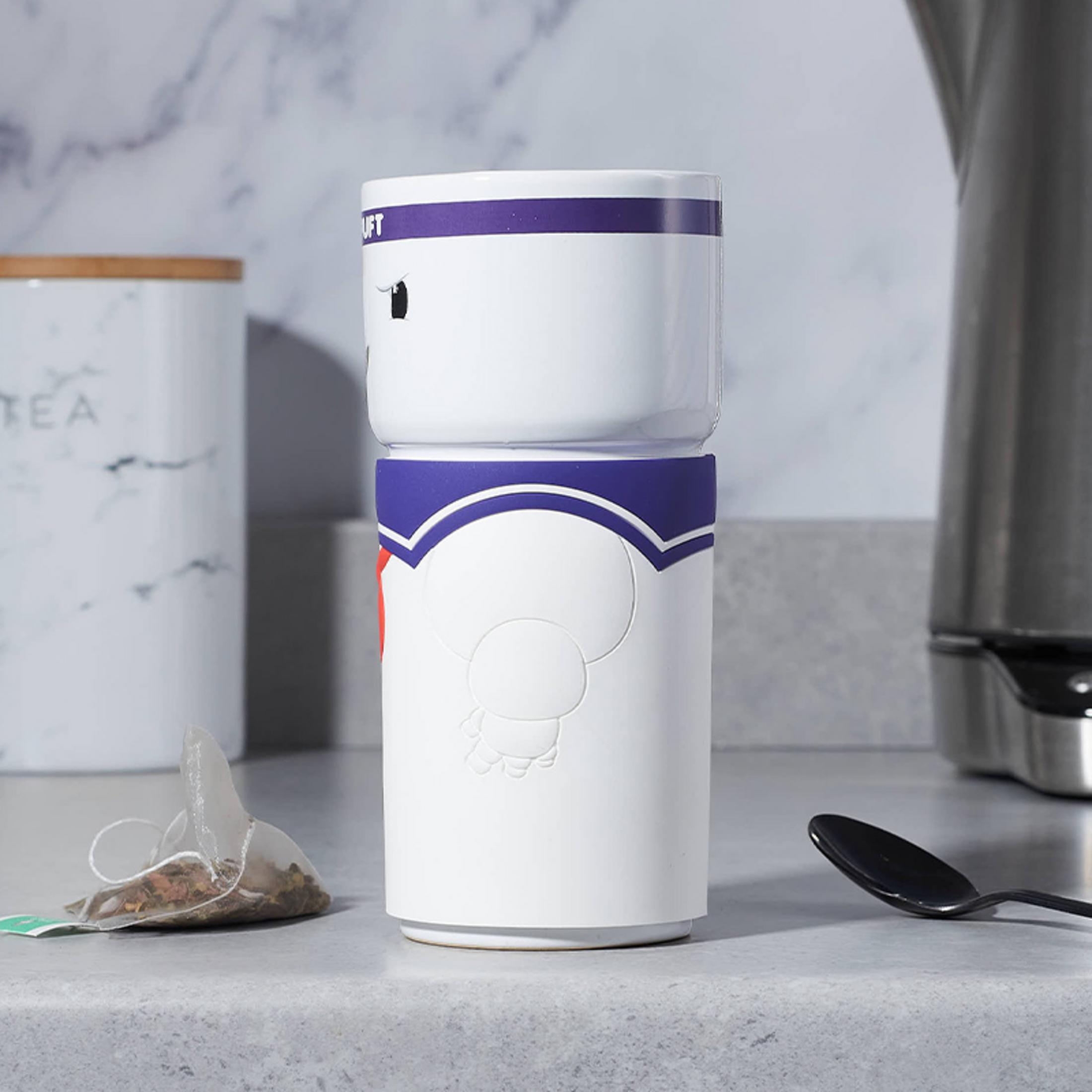 Weißer Becher mit Marshmallow-Design. Teebeutel und Löffel befinden sich in der Nähe des Bechers auf einer grauen Theke. Küchenumgebung.