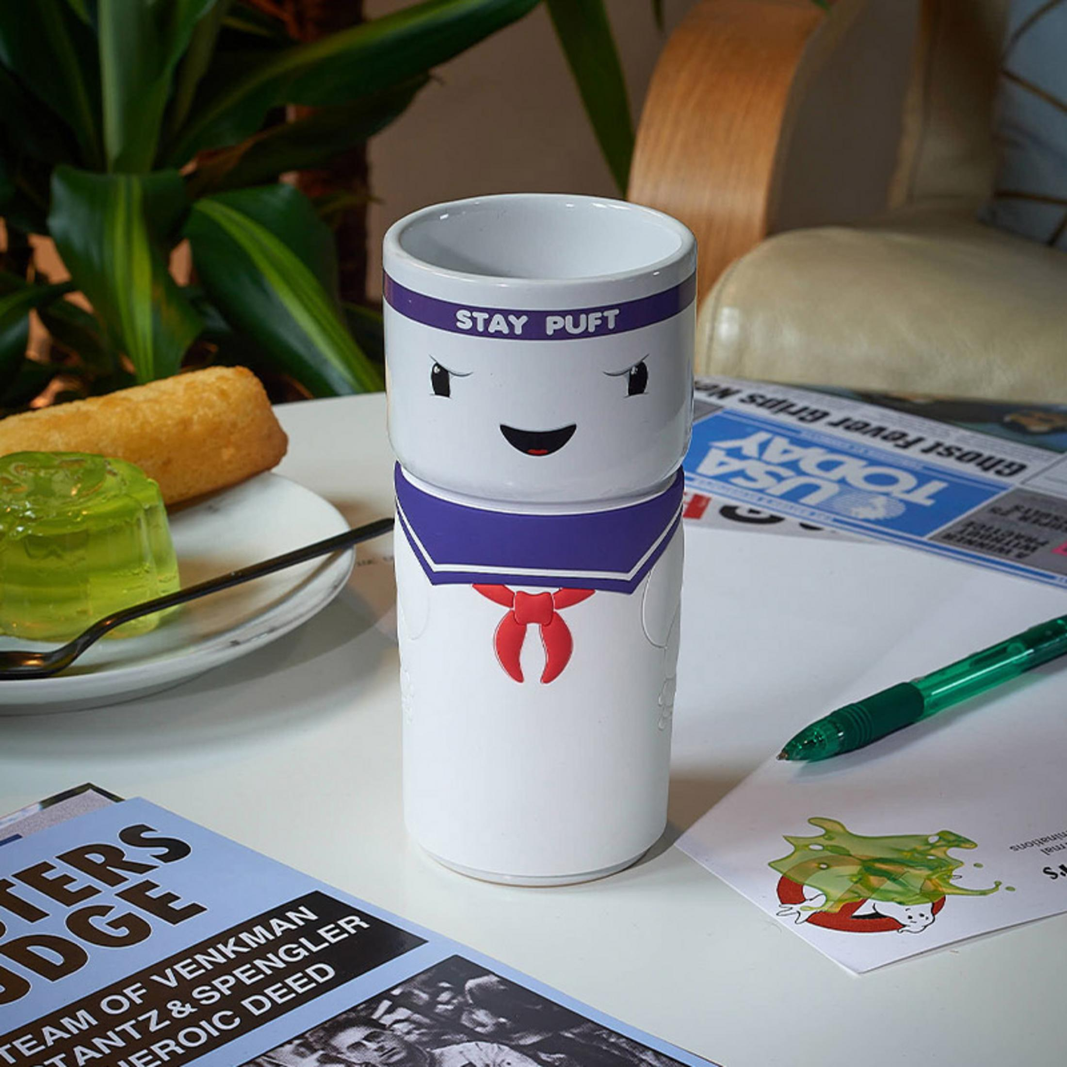 Ein weißer Stay Puft Marshmallow Man Becher steht auf einem Tisch mit einem grünen Stift und einer Zeitung.