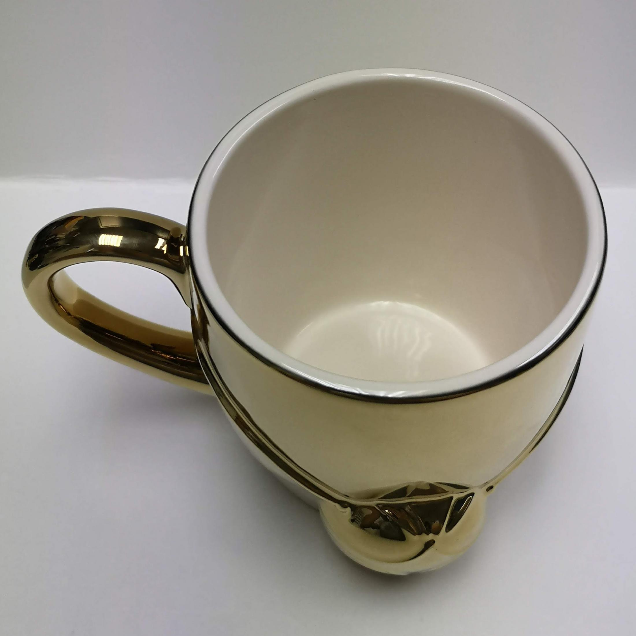 Goldfarbener Becher mit Henkel und Design am Boden, weißer Hintergrund.