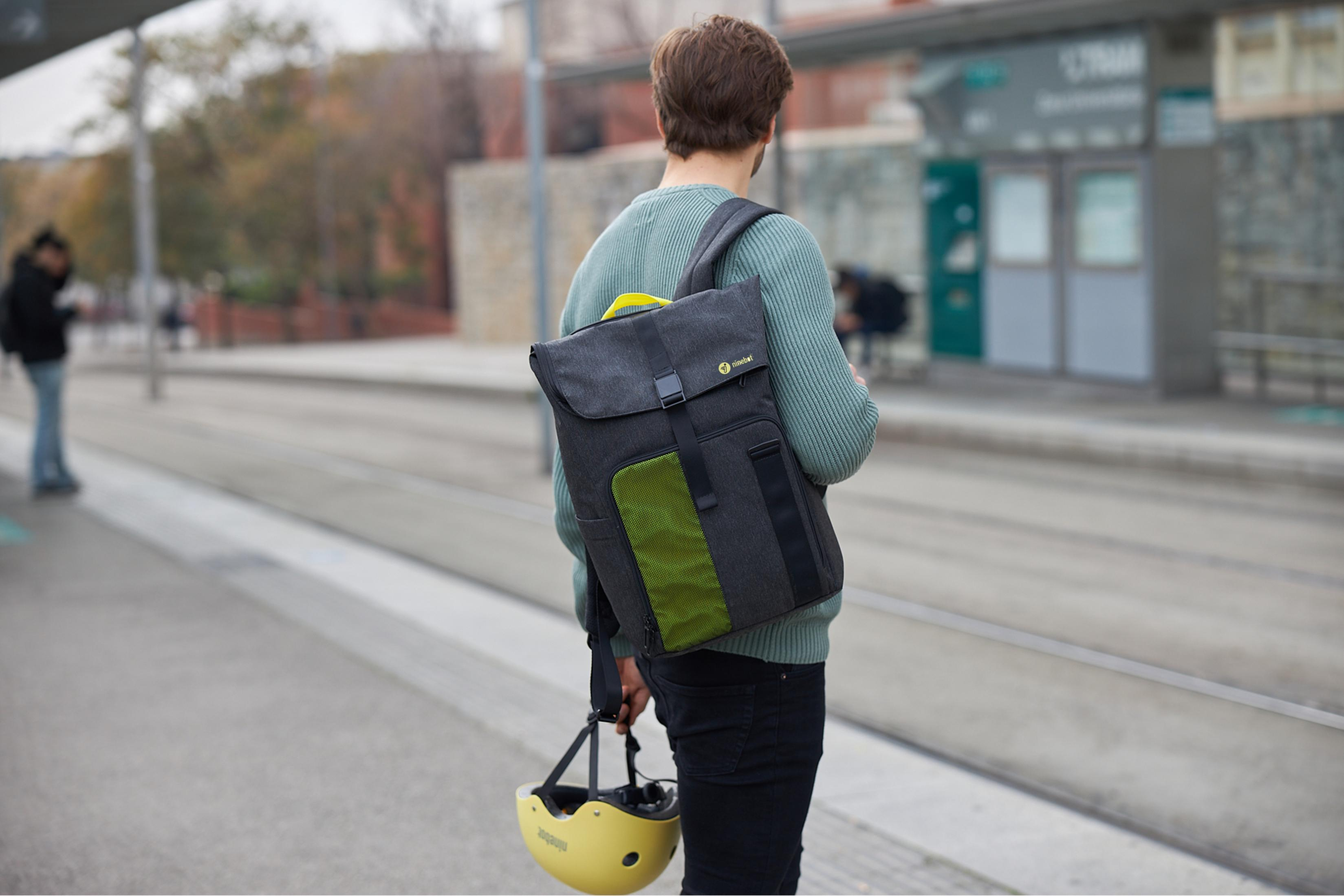 Ein Mann geht mit Rucksack und Helm in der Nähe einer Straßenbahnhaltestelle. Gebäude im Hintergrund.