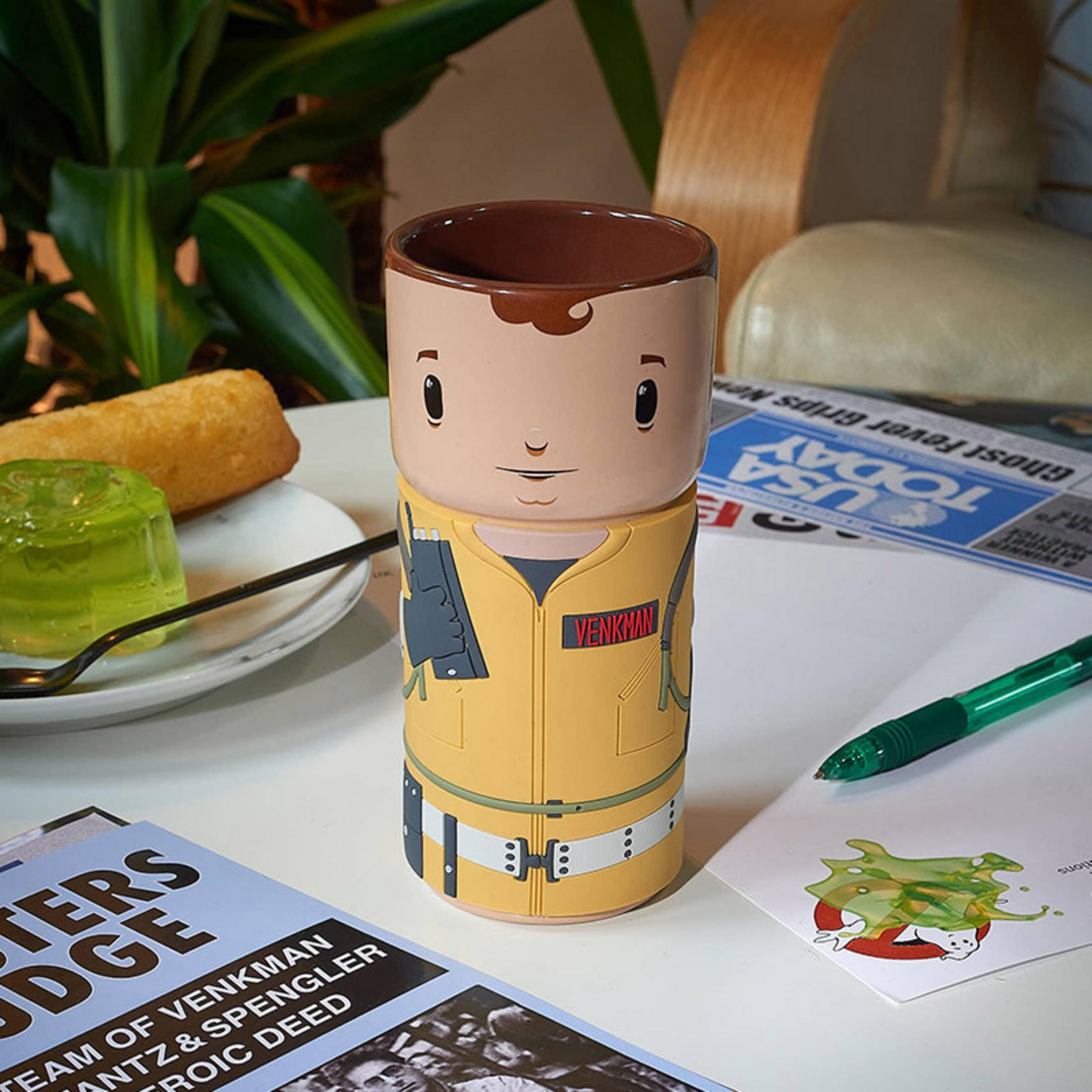 Ein Ghostbusters-Becher mit Venkman-Design, ein Snack und ein grünes Gelee auf einem Tisch.
