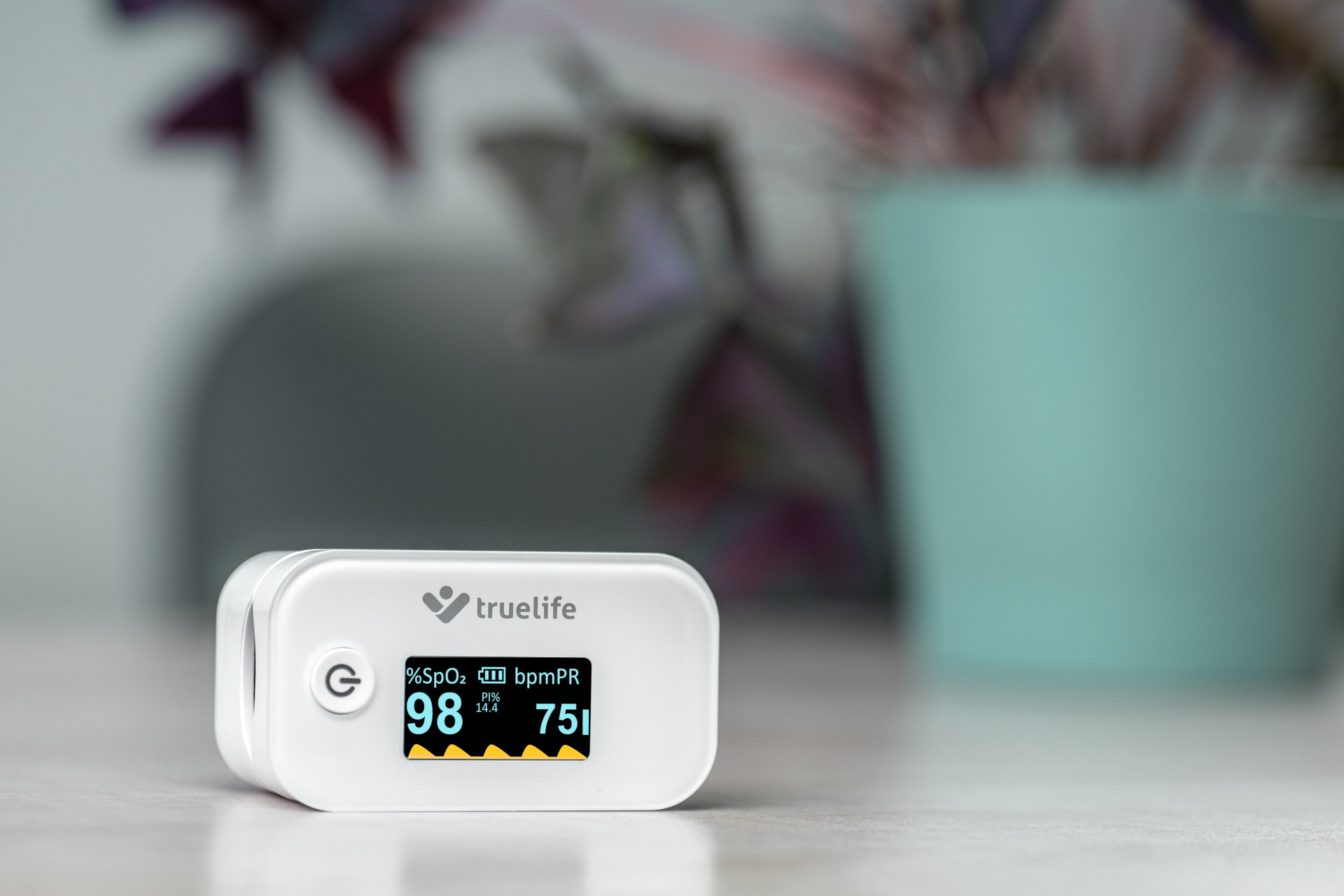 Weißes Pulsoximeter mit Digitalanzeige. Pflanze und Topf im Hintergrund.