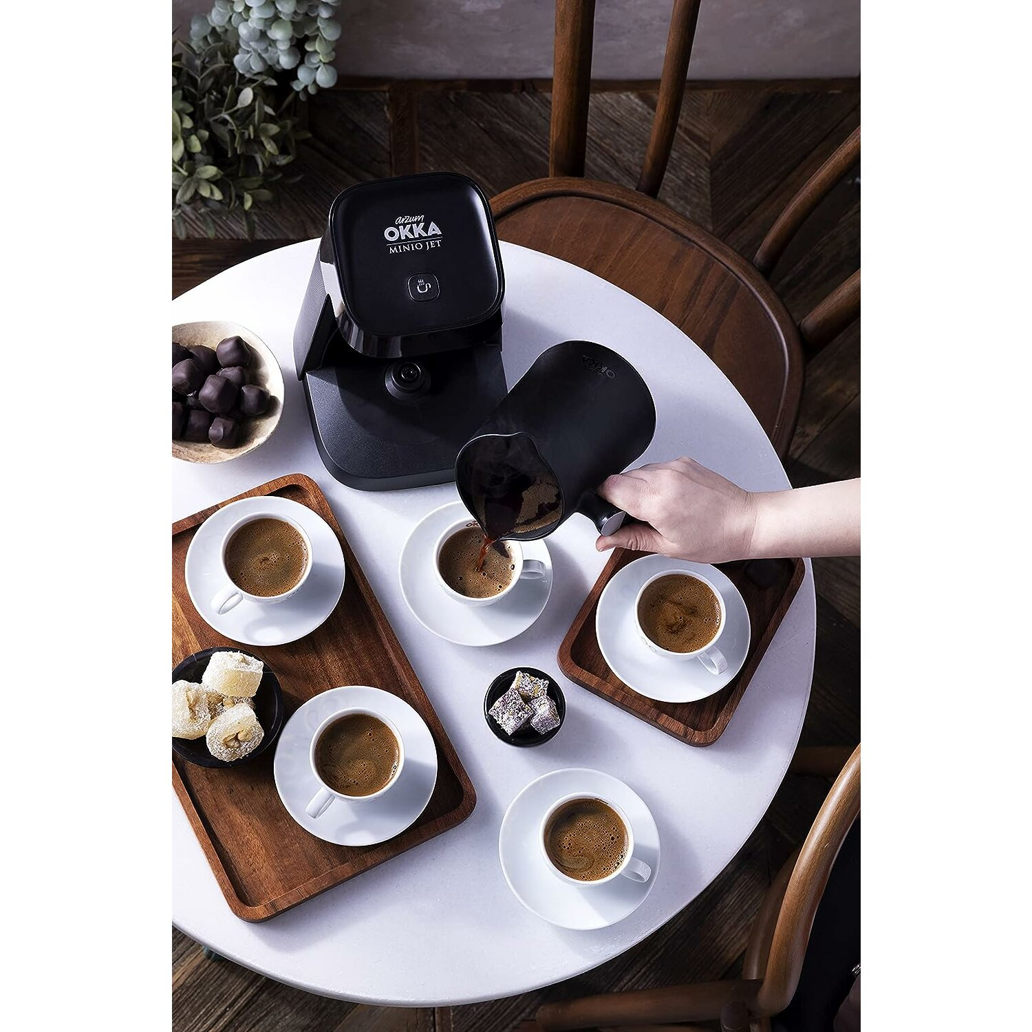 Eine Person gießt Kaffee aus einer schwarzen Kanne in kleine weiße Tassen auf einem weißen Tisch.