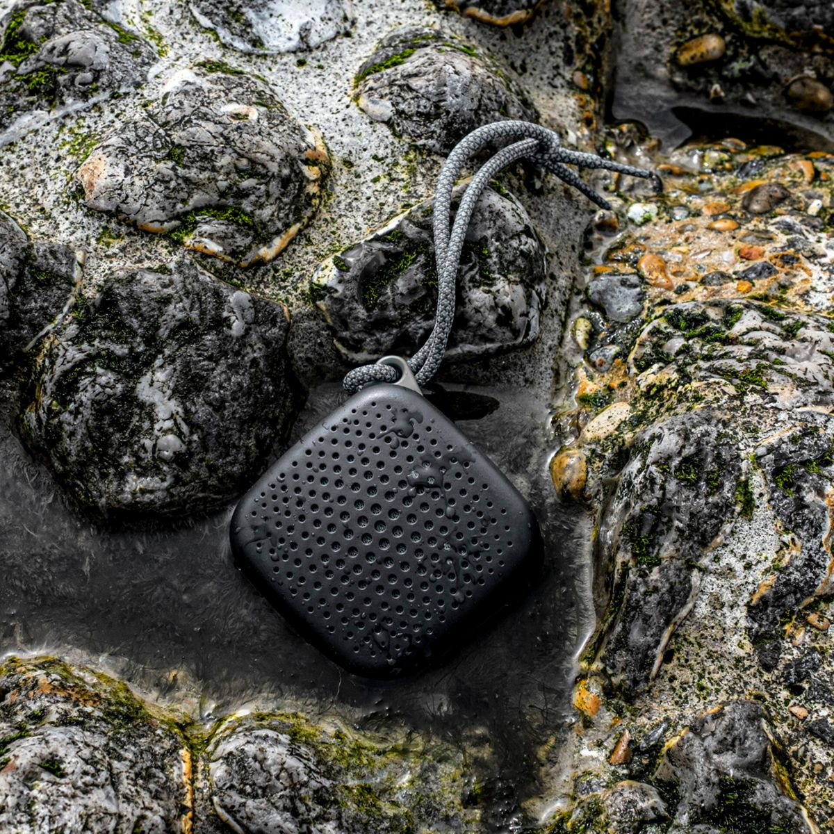 Czarny, kwadratowy głośnik na skałach w wodzie. Szary sznurek jest przymocowany do góry.