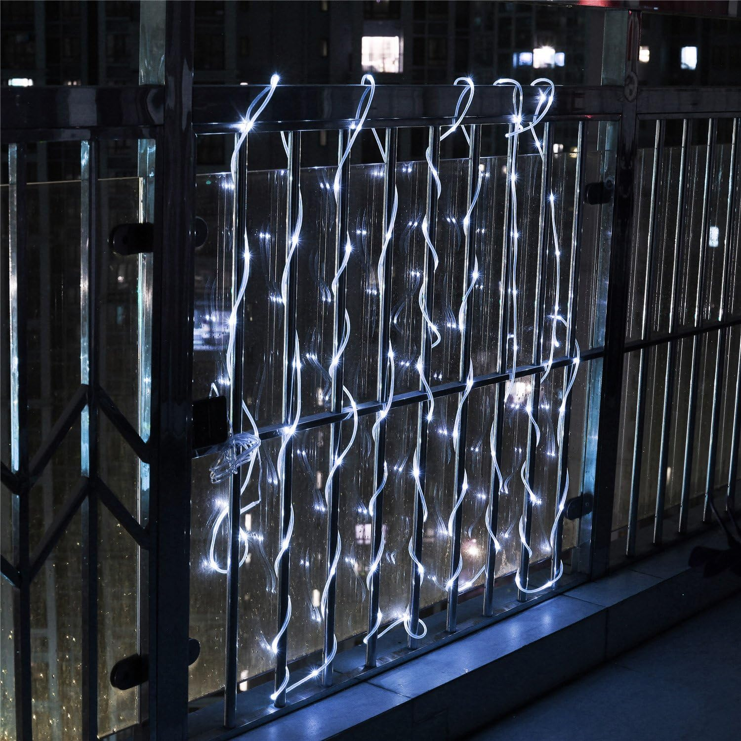 Weiße Lichterketten hängen nachts an einem Metallgeländer und reflektieren das Stadtlicht. Im Hintergrund sind Gebäude zu sehen.