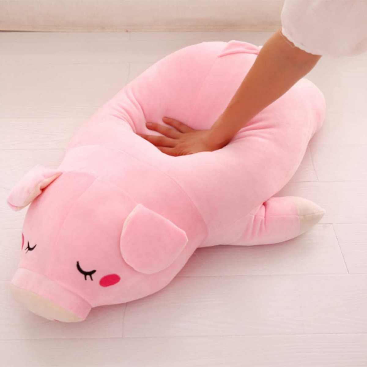 Ein rosa, schweineförmiges Plüschkissen, in das eine Hand drückt. Das Schwein hat geschlossene Augen und Rouge.
