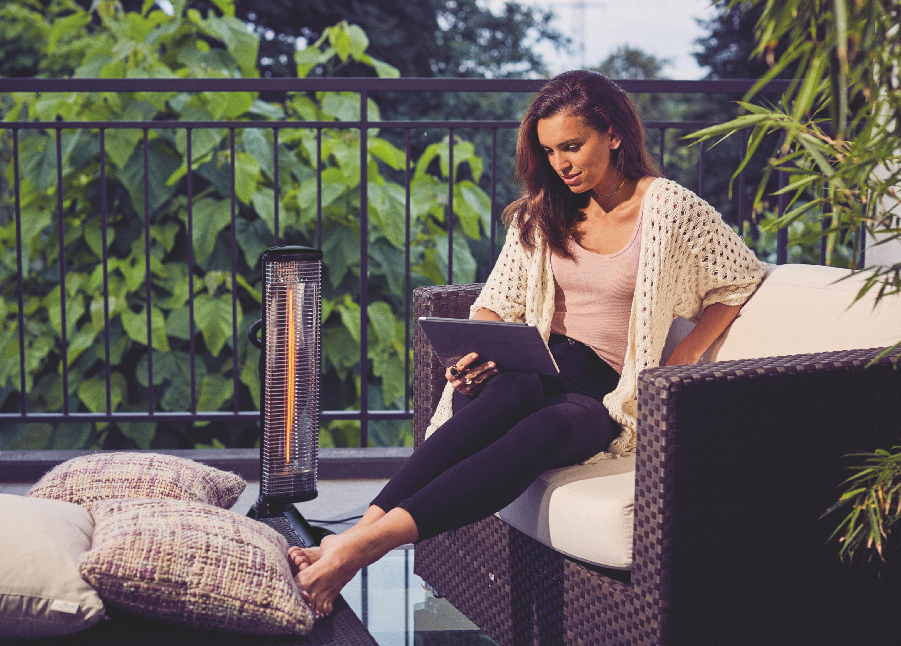 Eine Frau sitzt auf einer Couch auf einer Terrasse und benutzt ein Tablet, mit Heizung und Pflanzen in der Nähe.