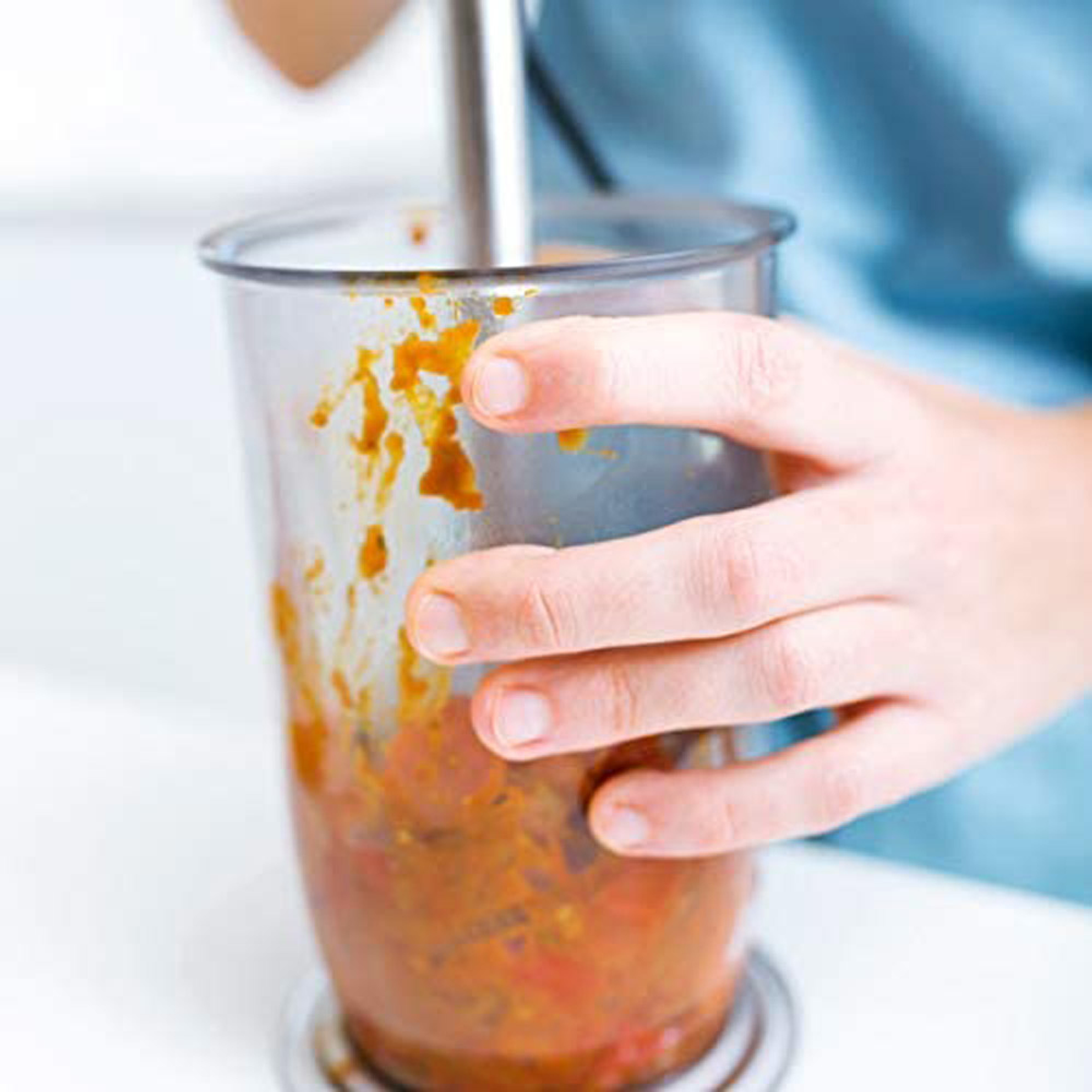 Eine Hand hält einen Stabmixer in einem Glas, der eine rötliche Mischung mixt. Weiße Arbeitsplatte.