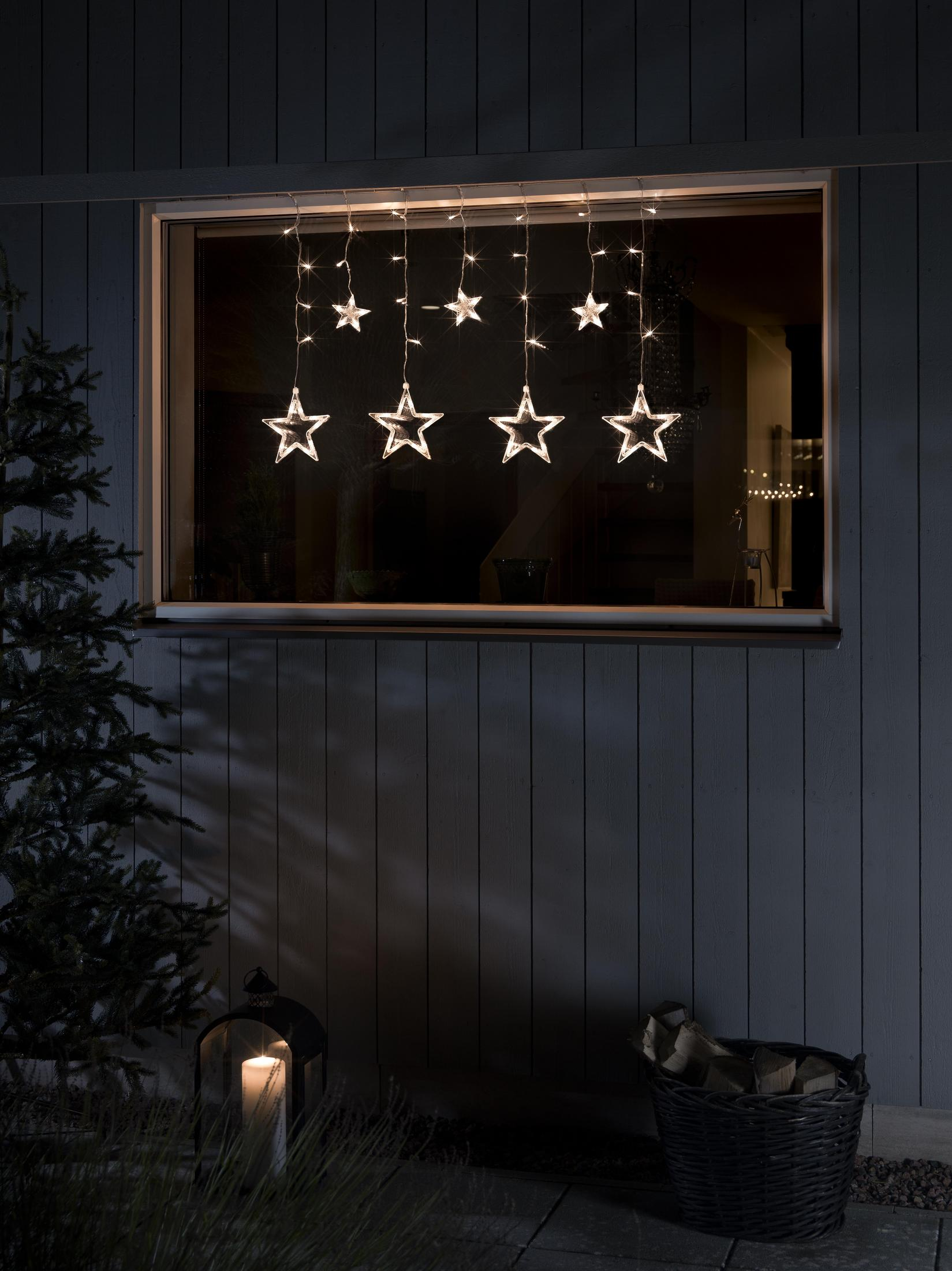 Fenster mit sternförmigen Lichtern. Eine brennende Kerze, ein Korb und ein Baum sind draußen.